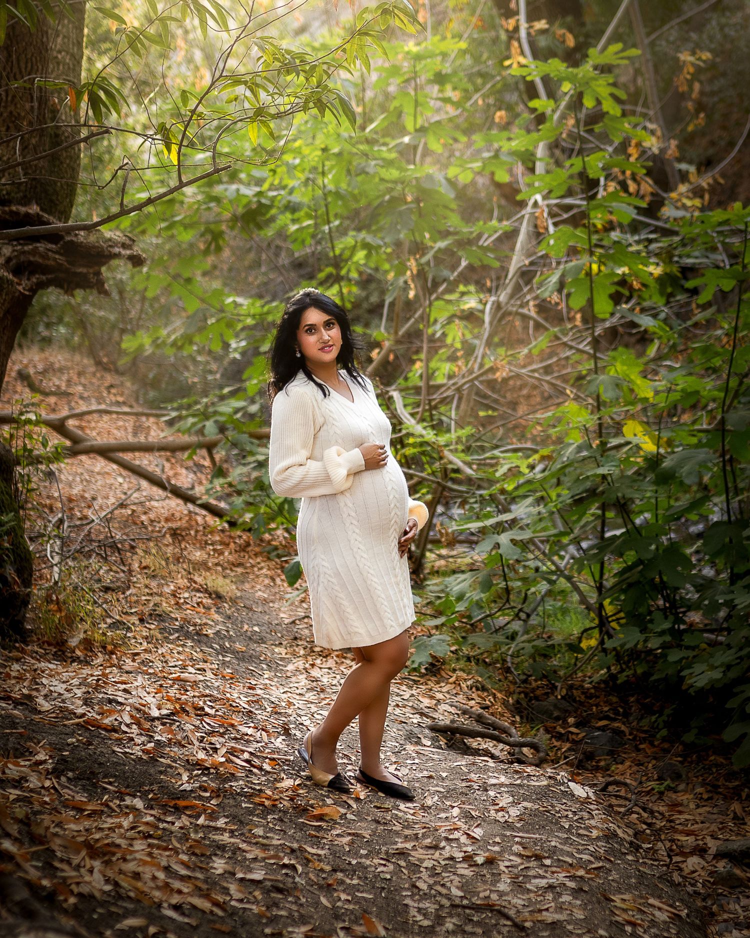 A person in a white dress poses on a wooded trail surrounded by lush green foliage and dappled sunlight.