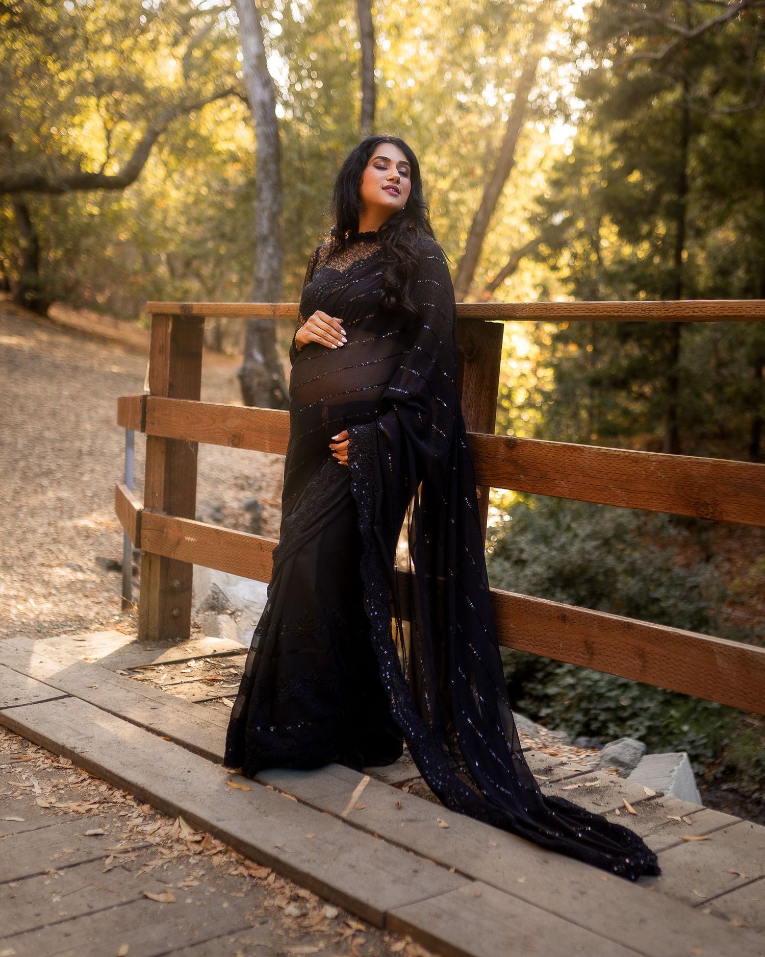 A figure in a flowing black dress poses gracefully on wooden steps in a sunlit forest setting.