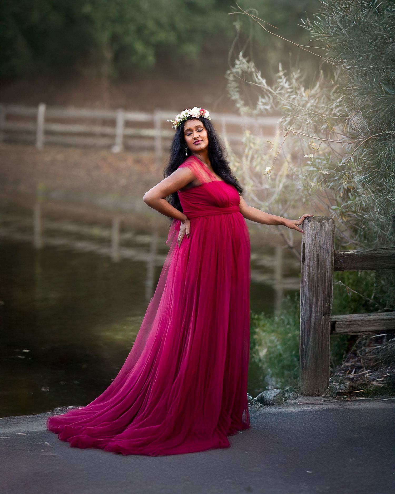 A flowing red tulle gown creates a dramatic silhouette against a rustic wooden fence near a tranquil pond.