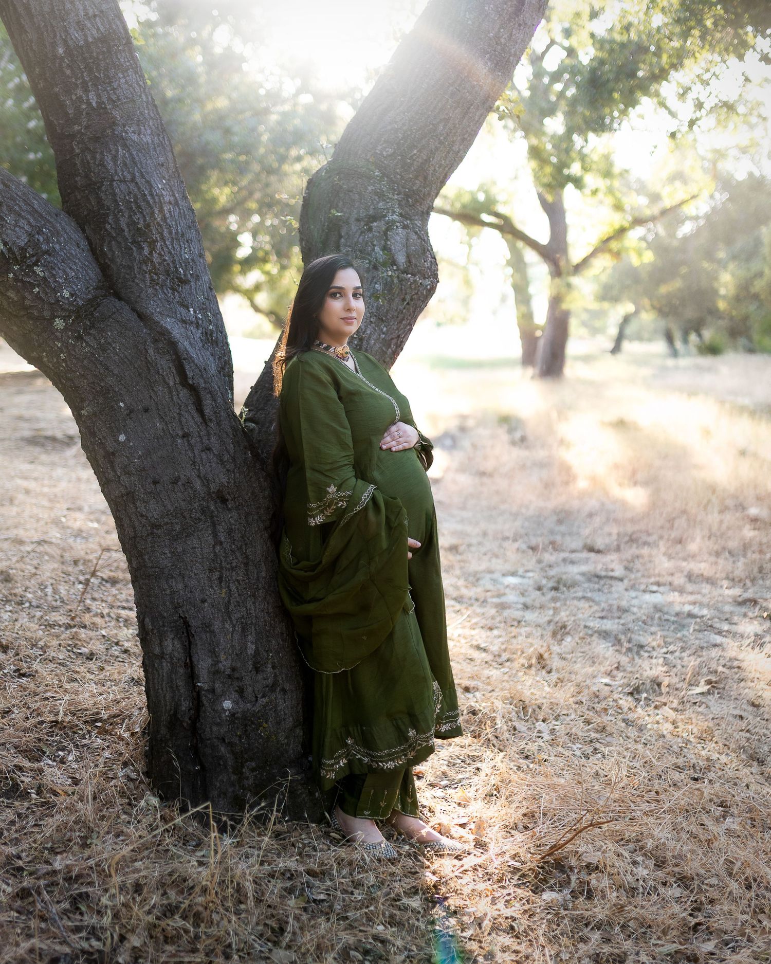A person in an olive green traditional dress stands gracefully against a large tree trunk in soft sunlit surroundings.
