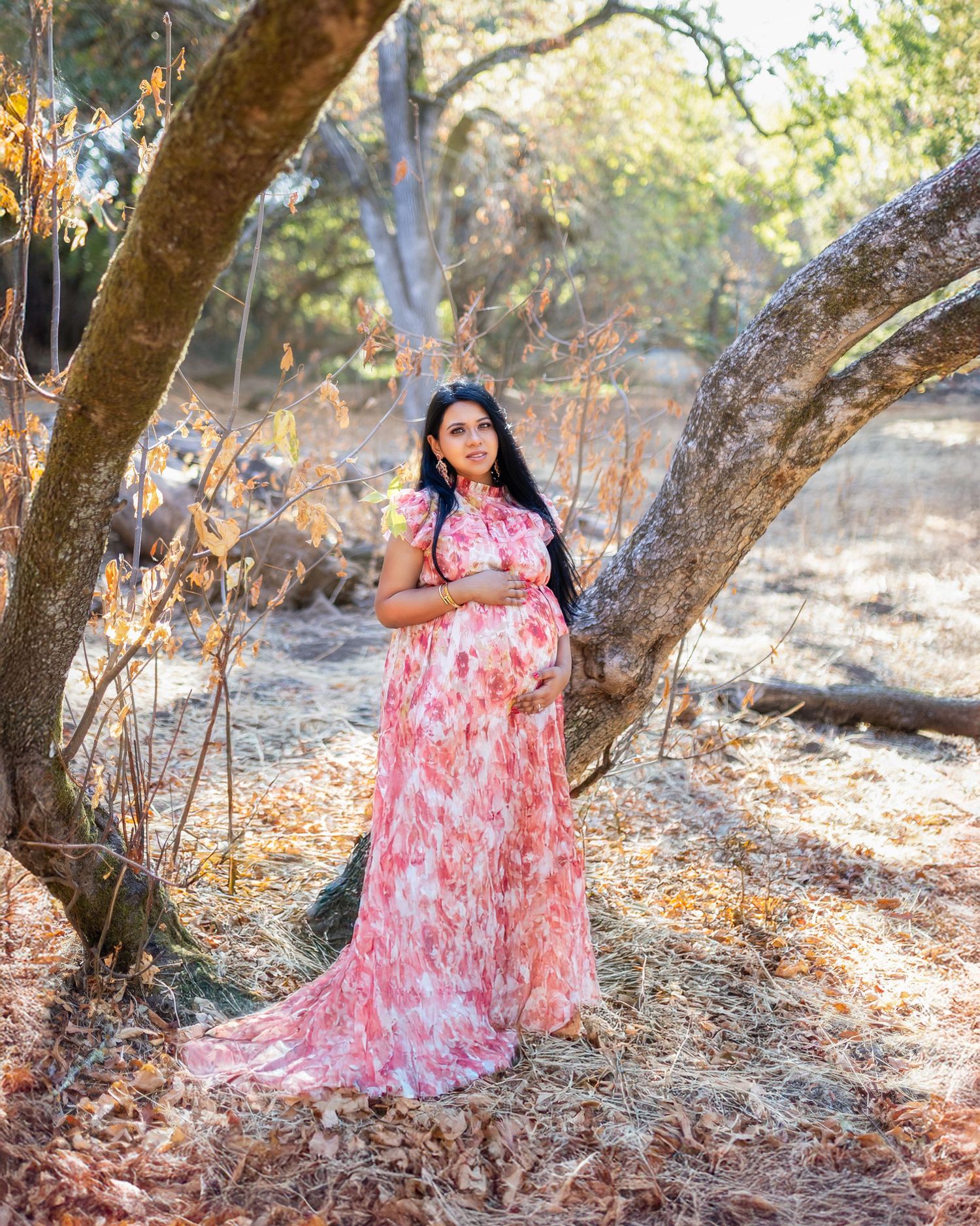 A person in a flowing pink and white floral dress poses gracefully against trees in a sunlit forest setting.