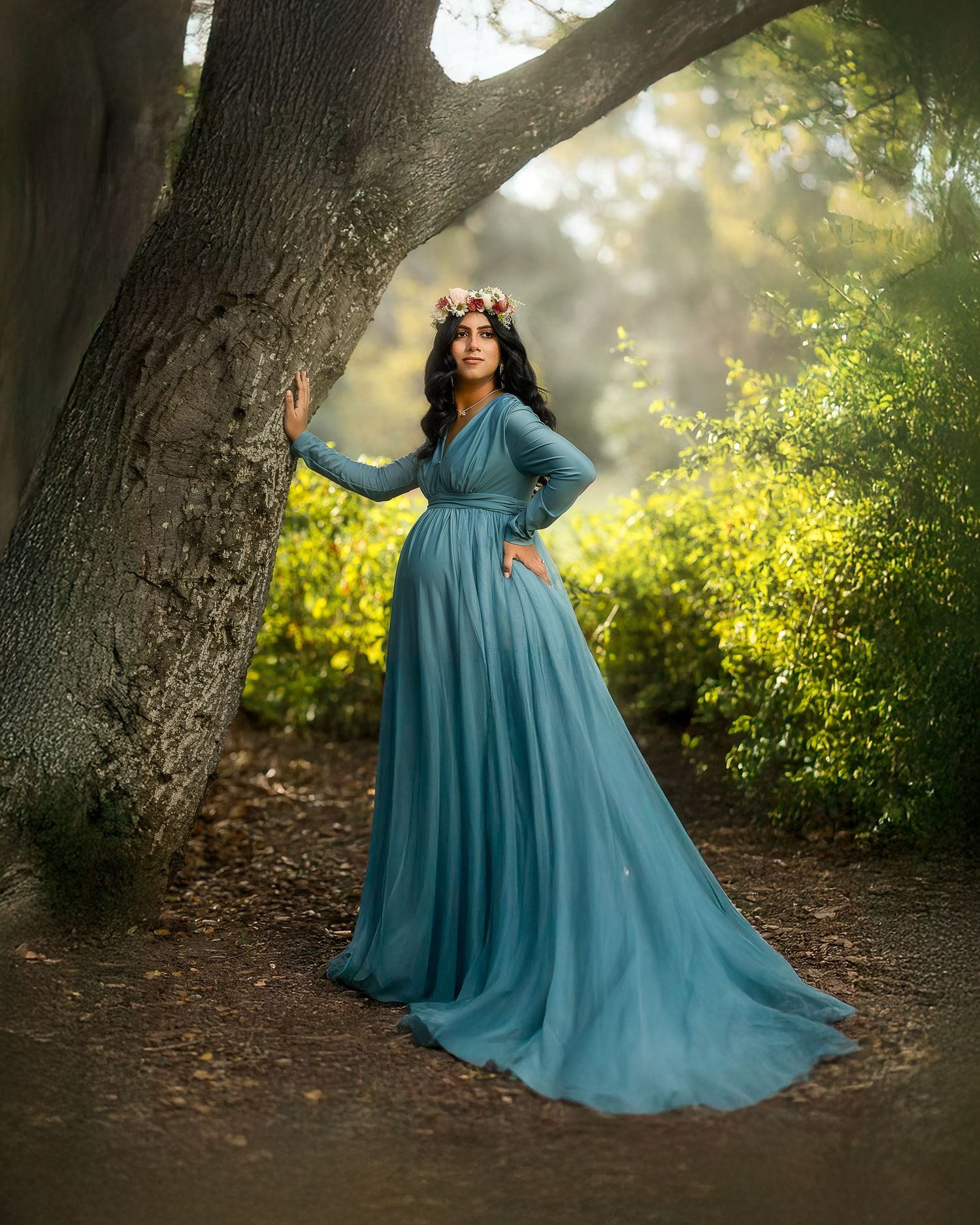 A flowing teal maternity gown catches sunlight beneath trees in a dreamy outdoor photoshoot setting.