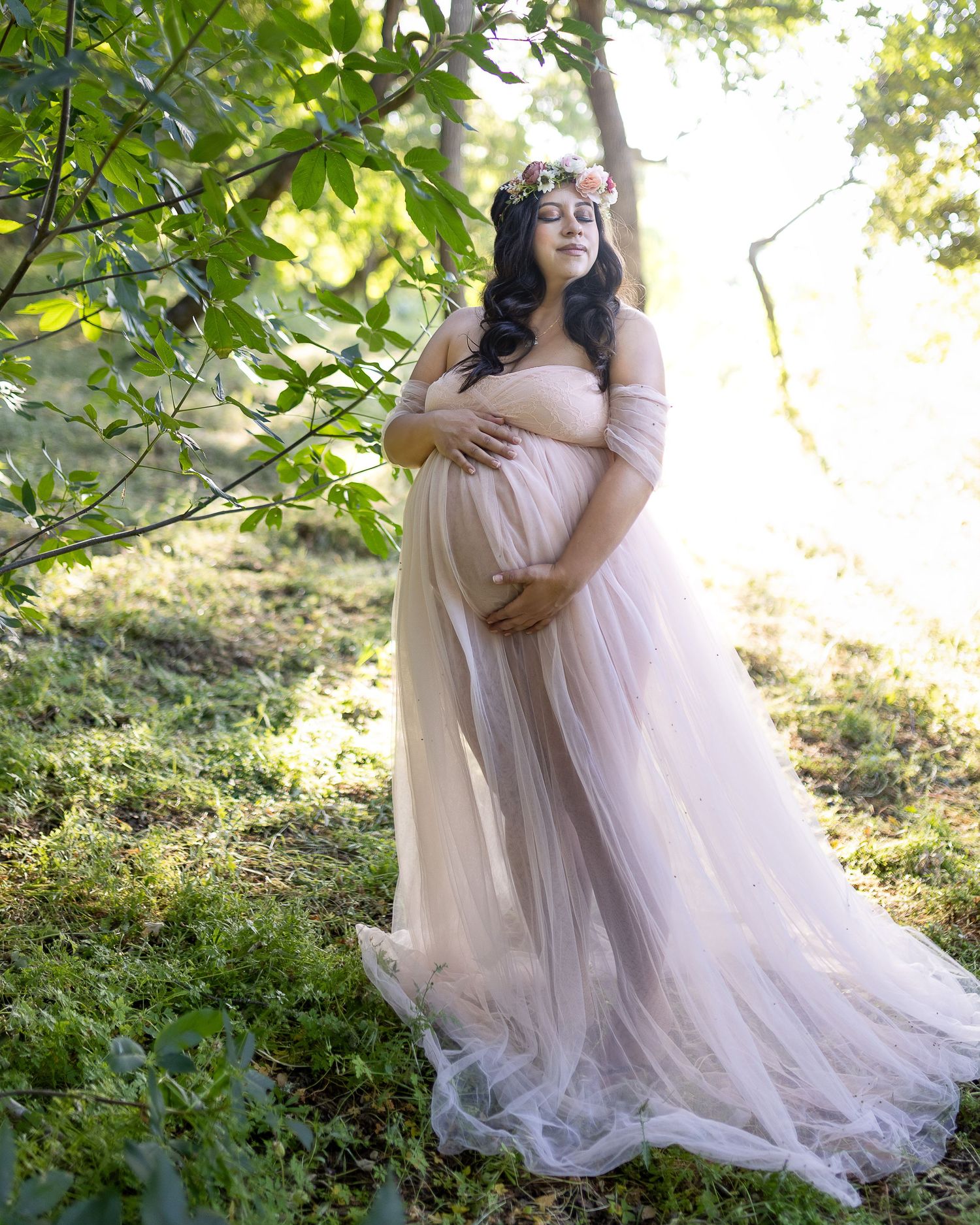 A romantic outdoor photoshoot features a flowing blush pink tulle gown against lush green foliage in natural sunlight.