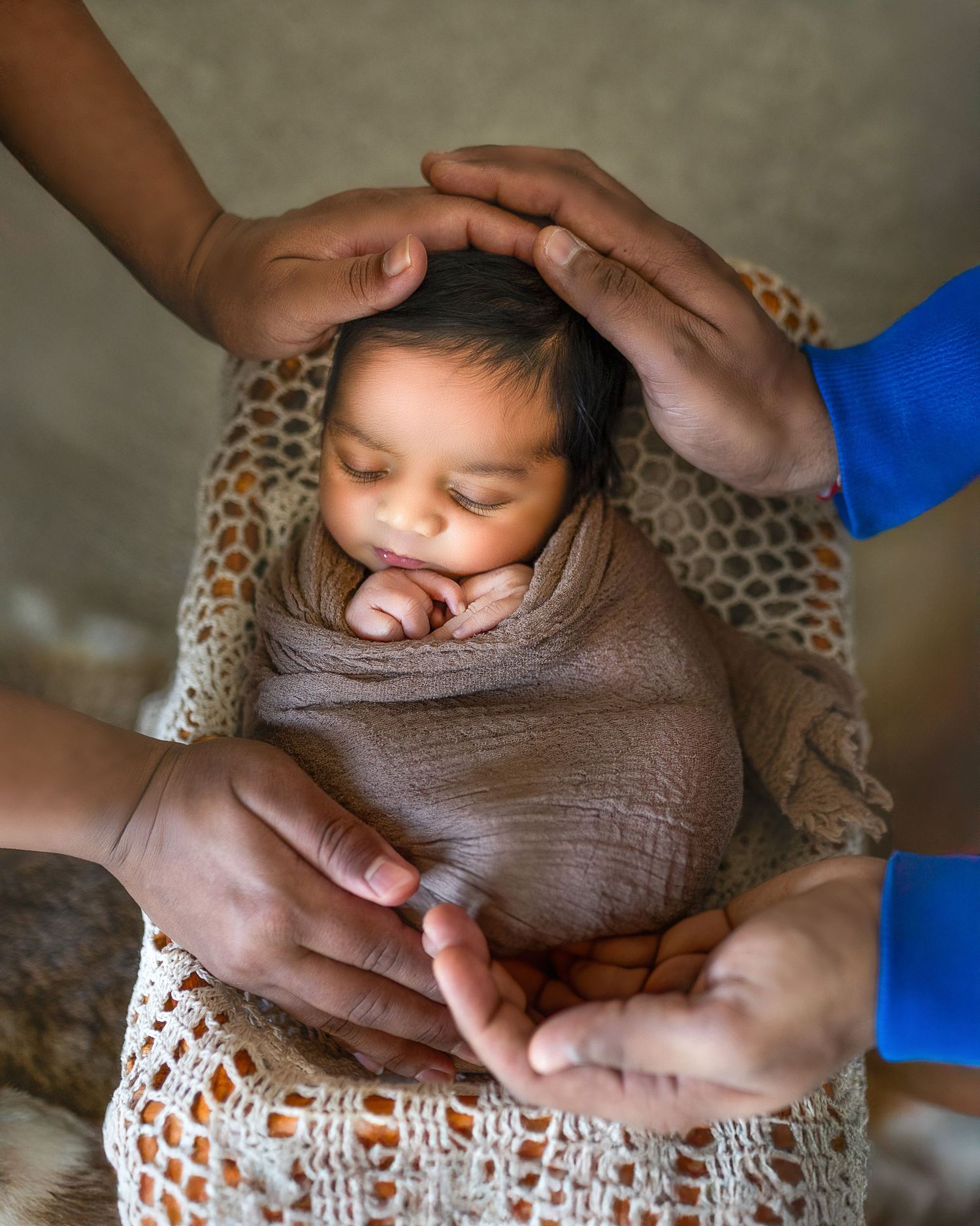 Gentle hands carefully wrap a sleeping newborn in a soft brown swaddle blanket during a photography session.