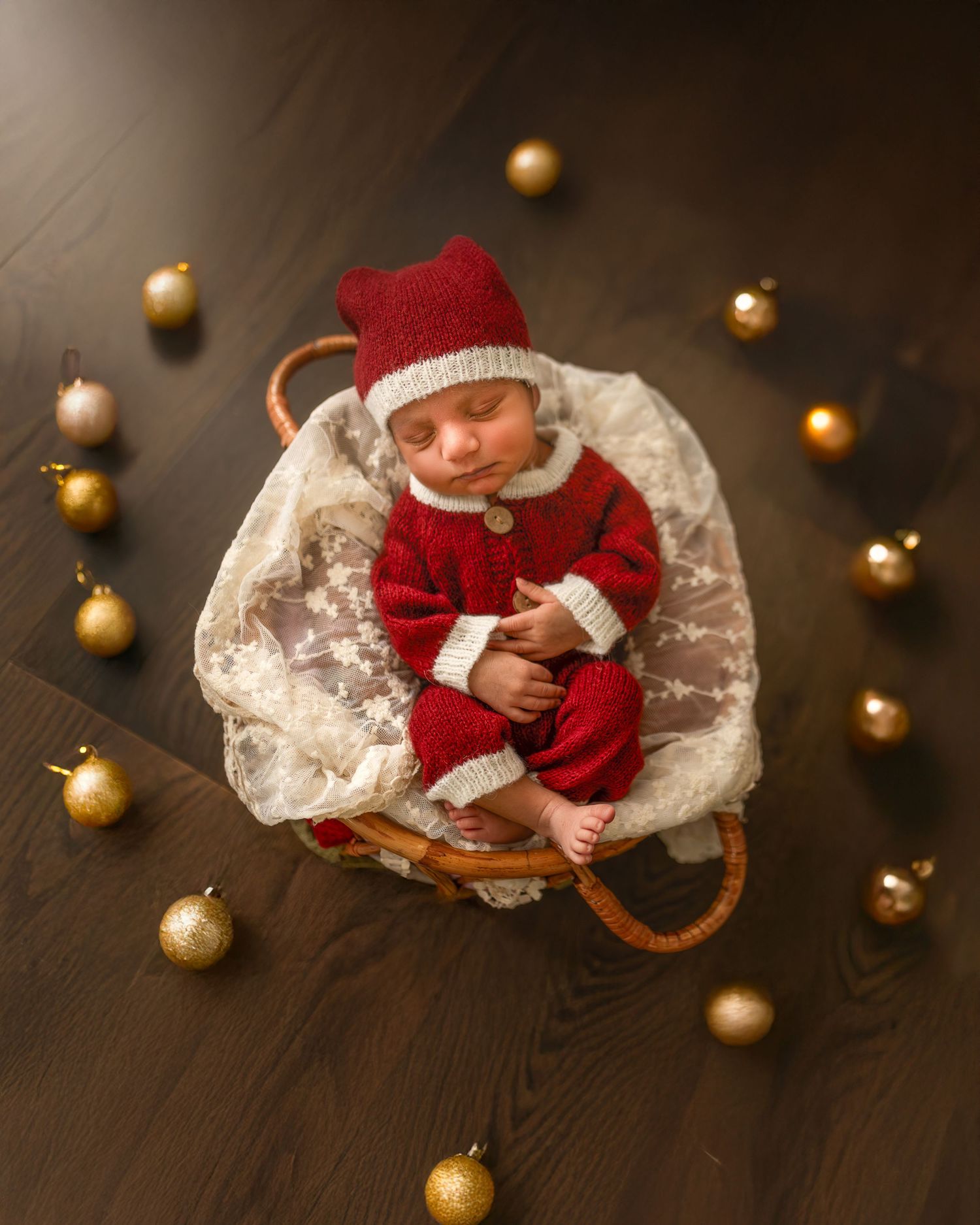 A sleeping newborn in red outfit rests in a woven basket surrounded by golden Christmas ornaments on dark wood floor.