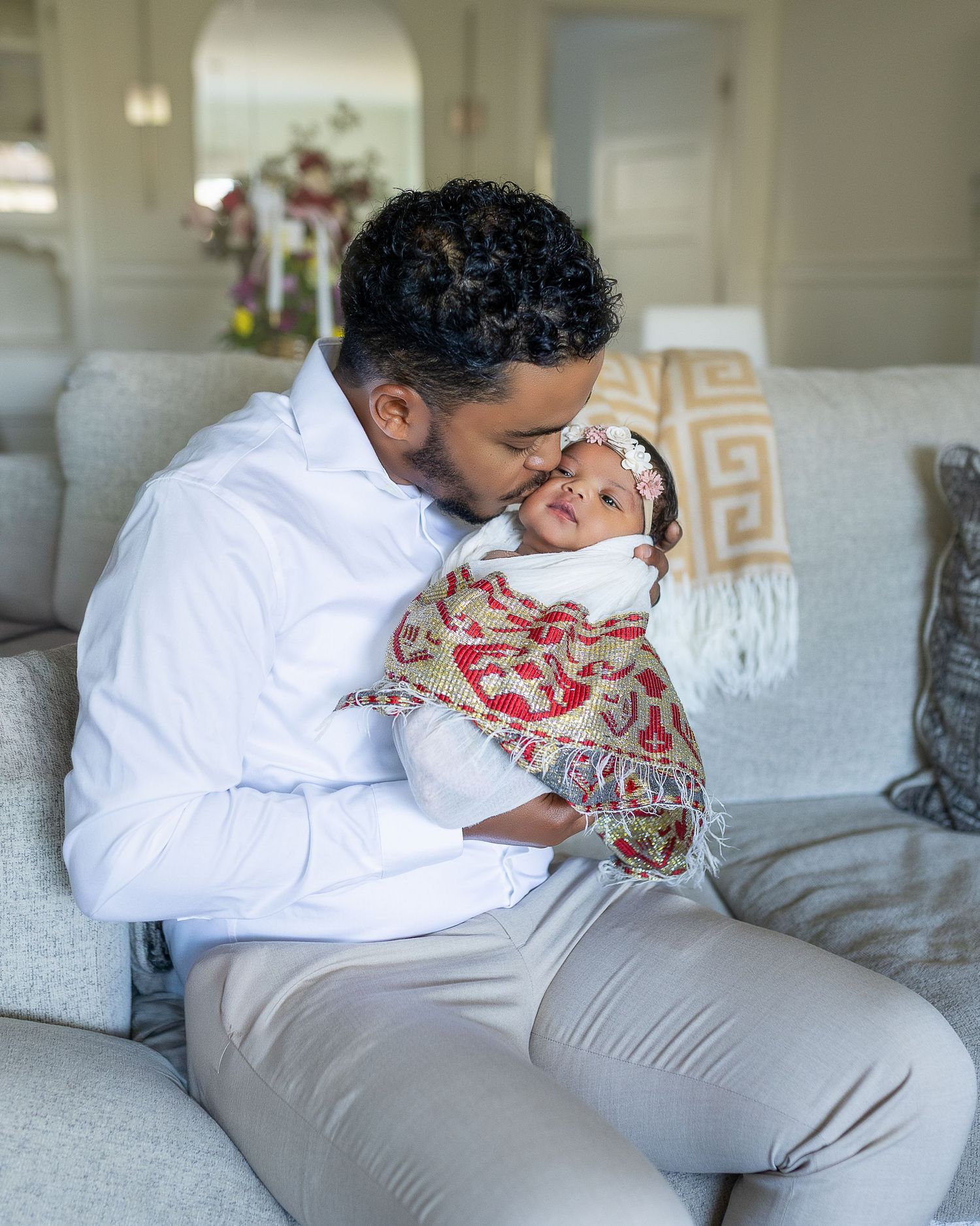 A tender moment shared between parent and swaddled newborn baby on a gray couch in a bright living room.
