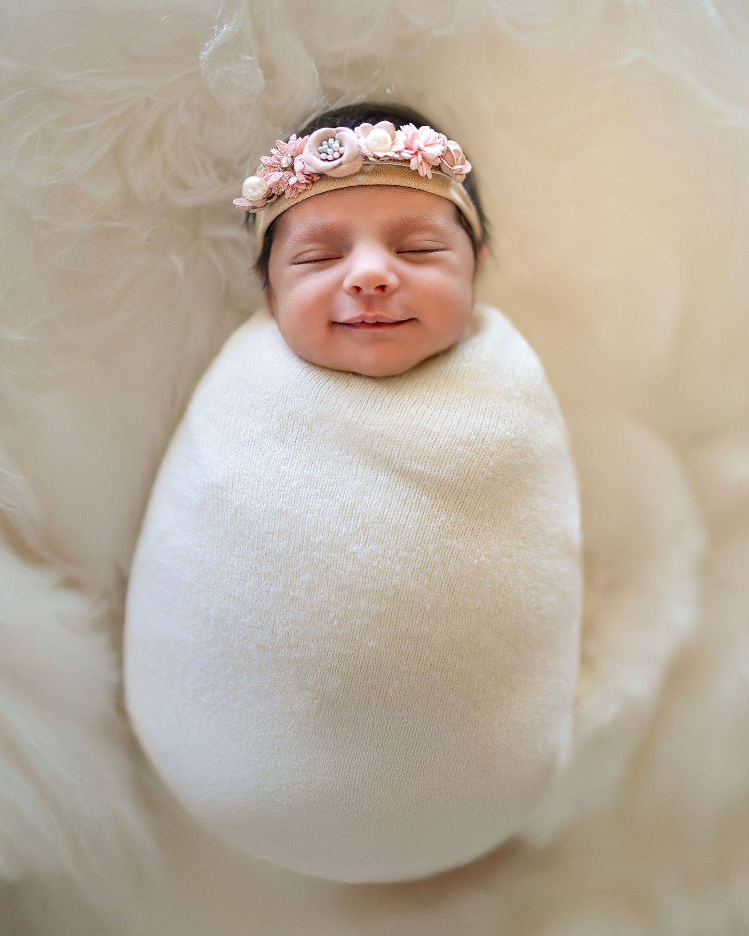 A sleeping newborn baby swaddled in white fabric with a delicate pink floral headband.