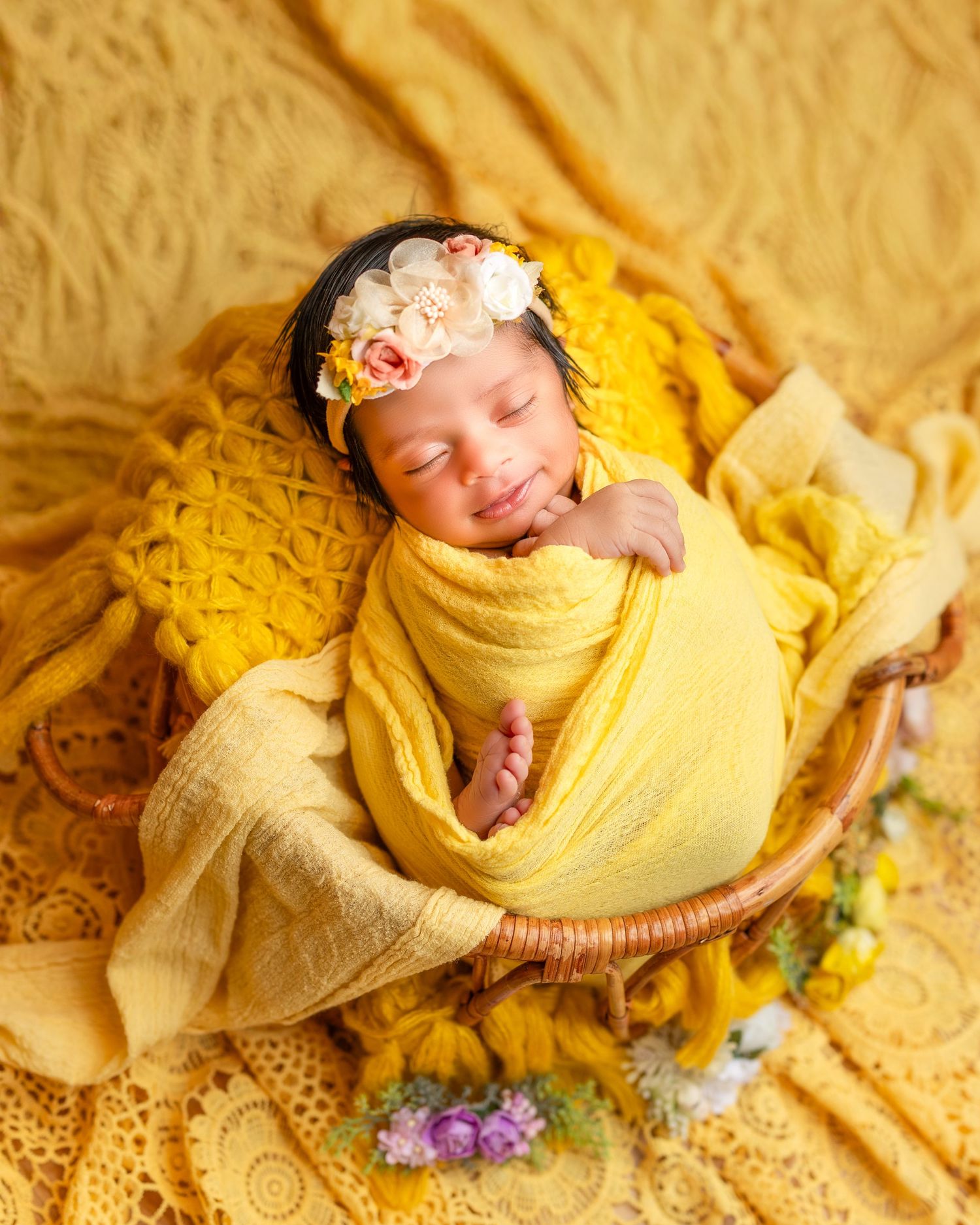 A sleeping newborn wrapped in yellow fabric with floral decorations and soft lighting creates a dreamy portrait setting.