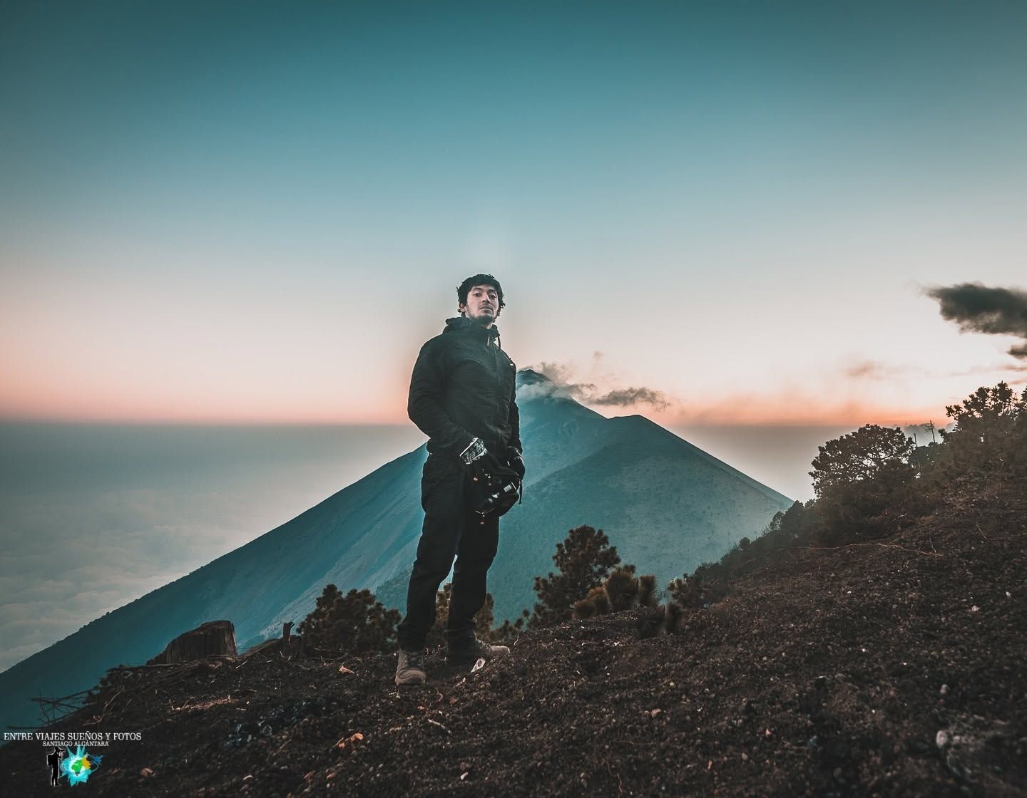 Person standing on volcanic hillside at sunset with erupting volcano and ocean in background.