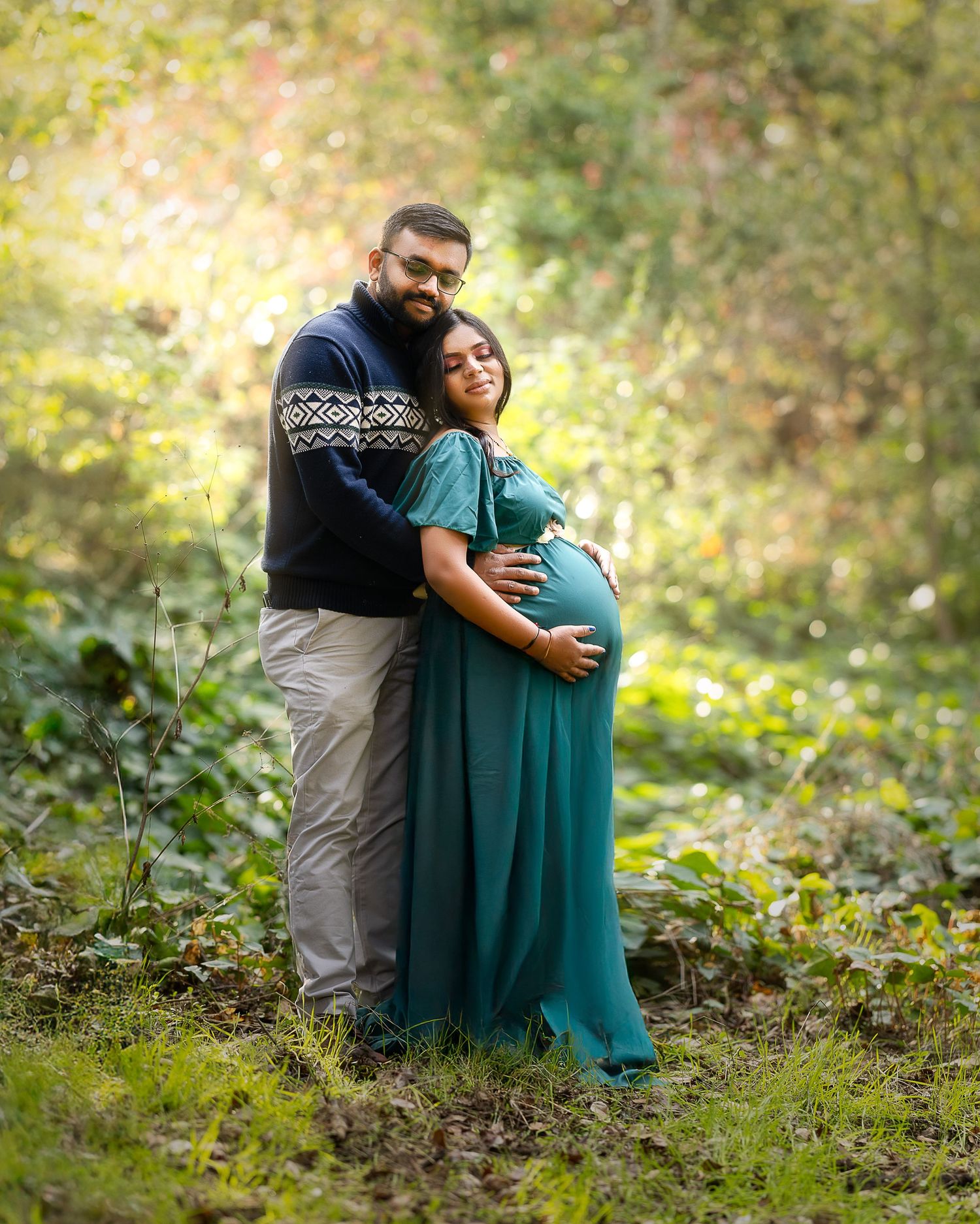 A couple embraces in a natural outdoor setting with sunlit greenery and foliage in the background.