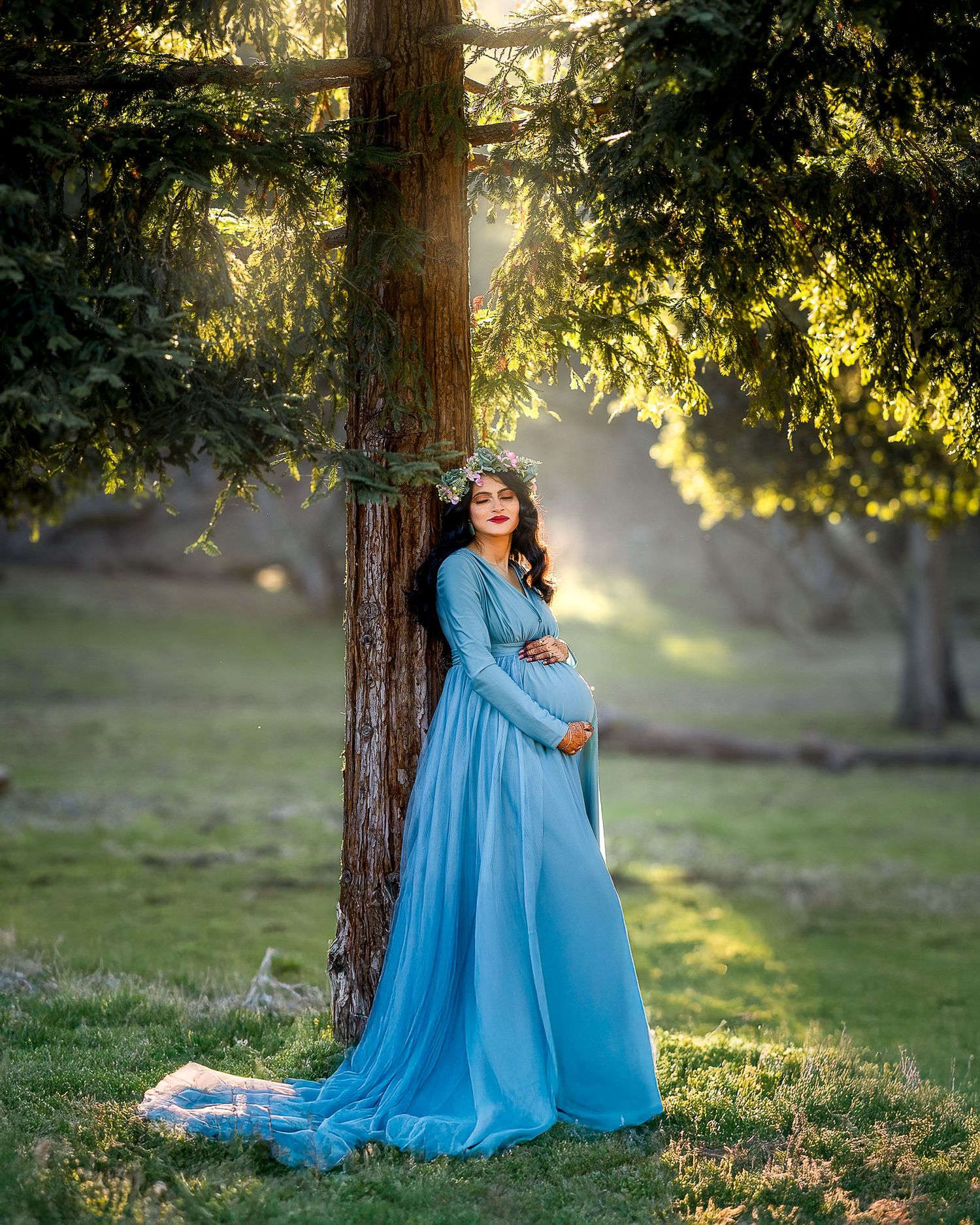 A flowing light blue maternity gown catches sunlight beneath tall trees in a dreamy outdoor setting.