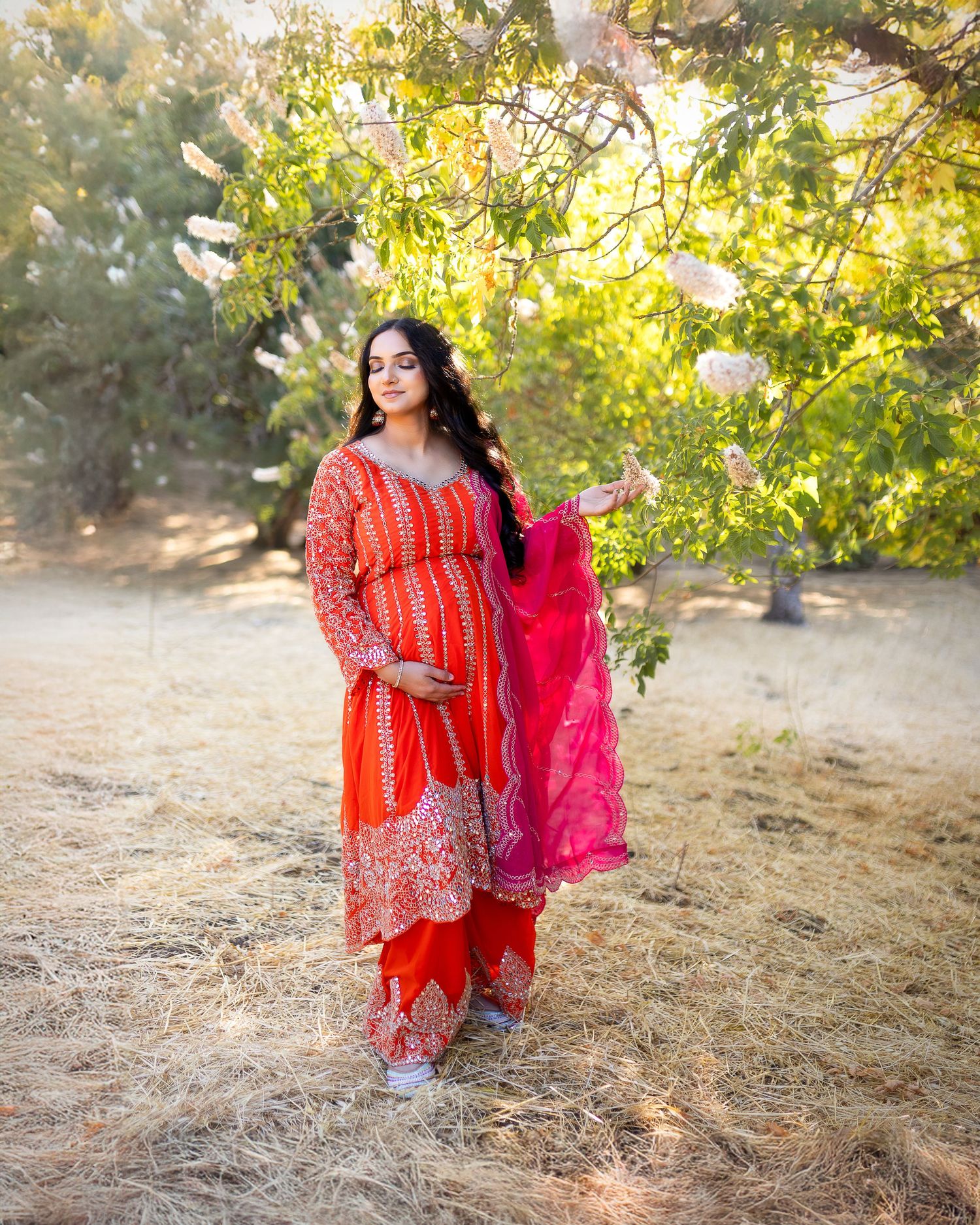 Traditional red and pink Indian dress flows gracefully in outdoor garden setting with golden sunlight filtering through trees.