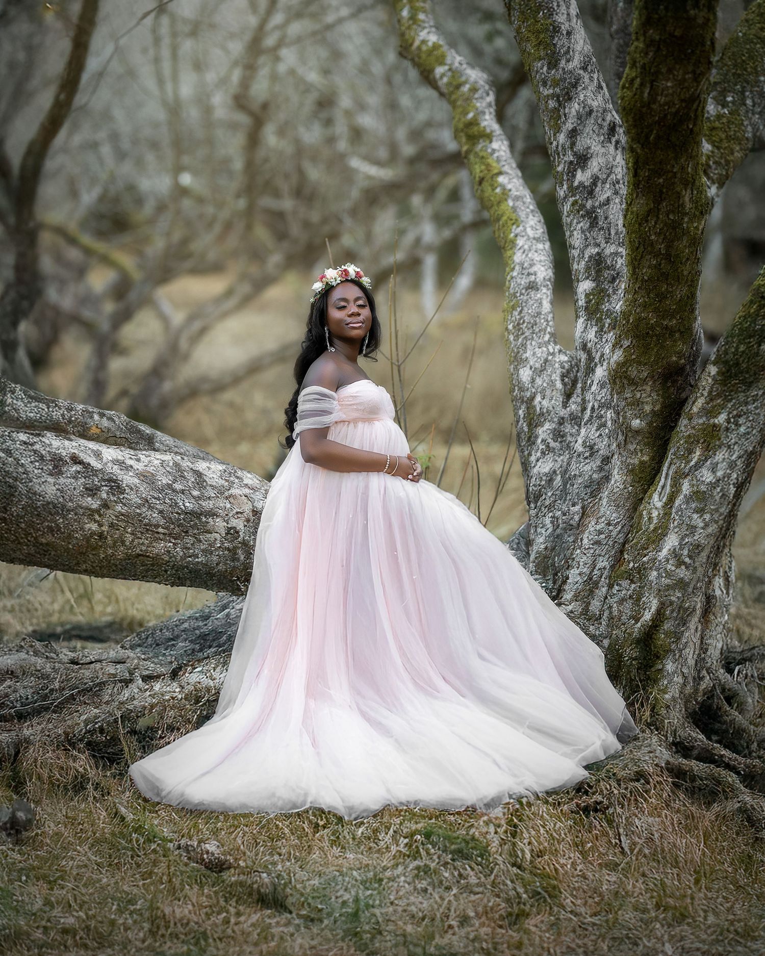 A person in a flowing pink tulle gown poses gracefully on a tree branch in a dreamy woodland setting.