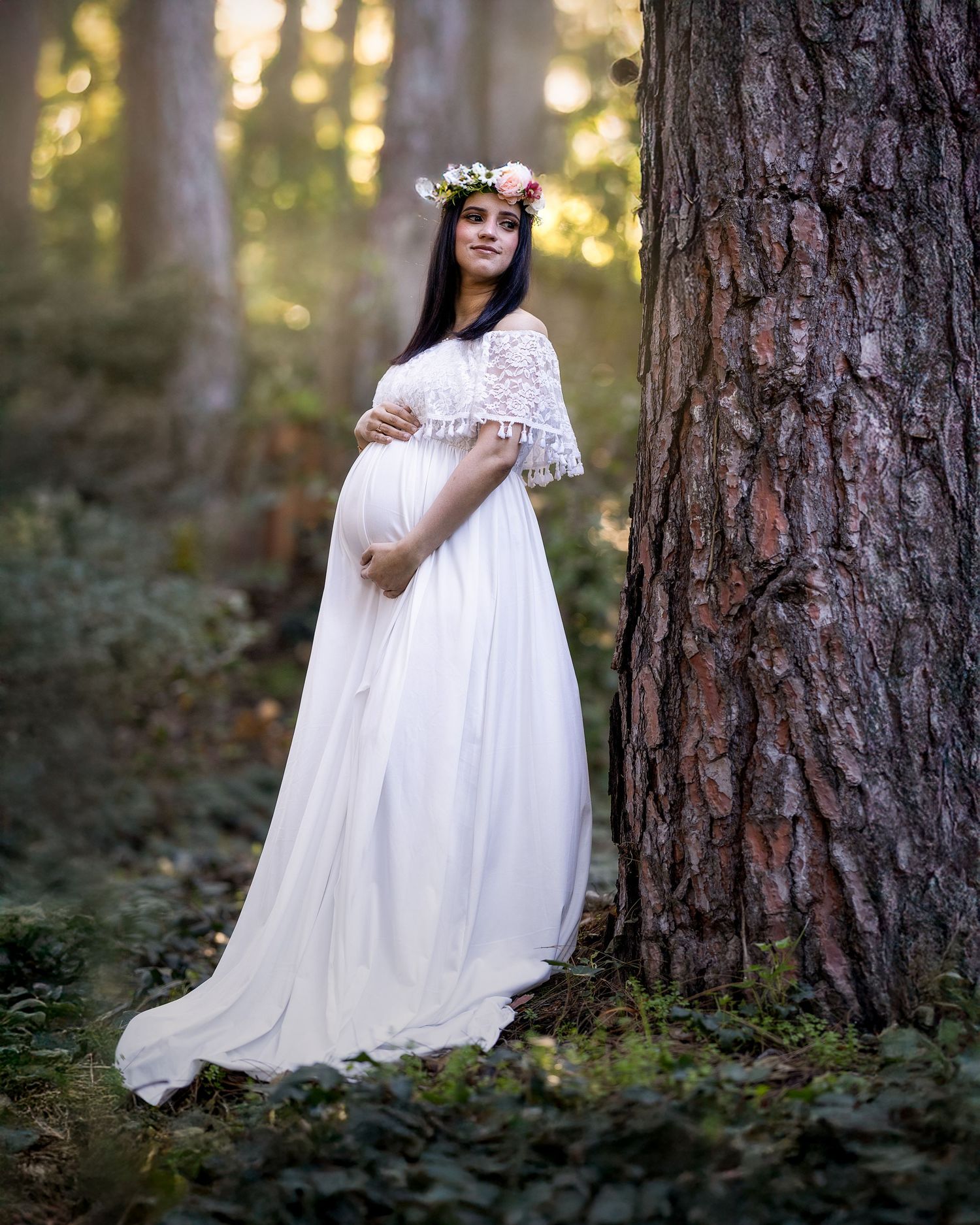 A dreamy maternity photo in a forest shows a woman in a flowing white lace dress and floral crown leaning against a large tree.