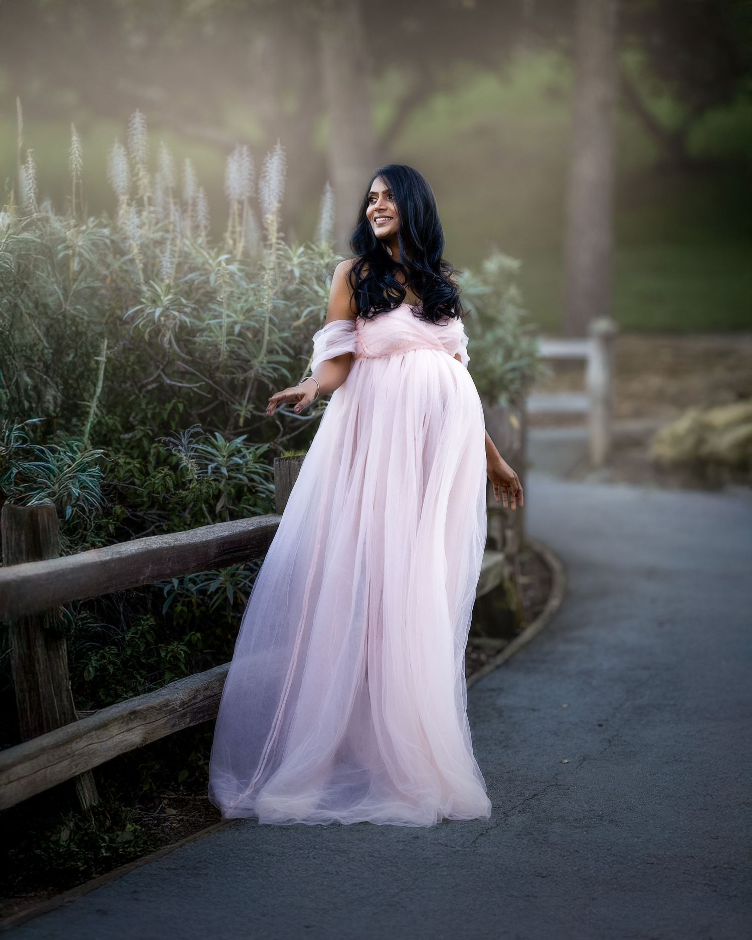 A person in a flowing pink tulle gown poses gracefully on a garden pathway in a dreamy outdoor setting.