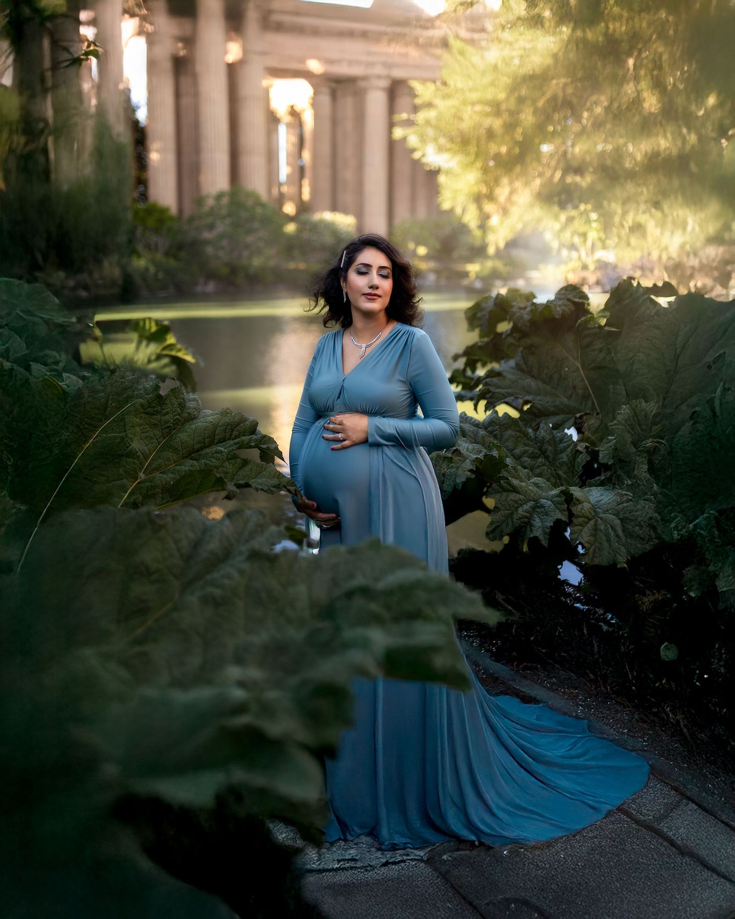 A person in a flowing blue dress poses gracefully among garden foliage at sunset.
