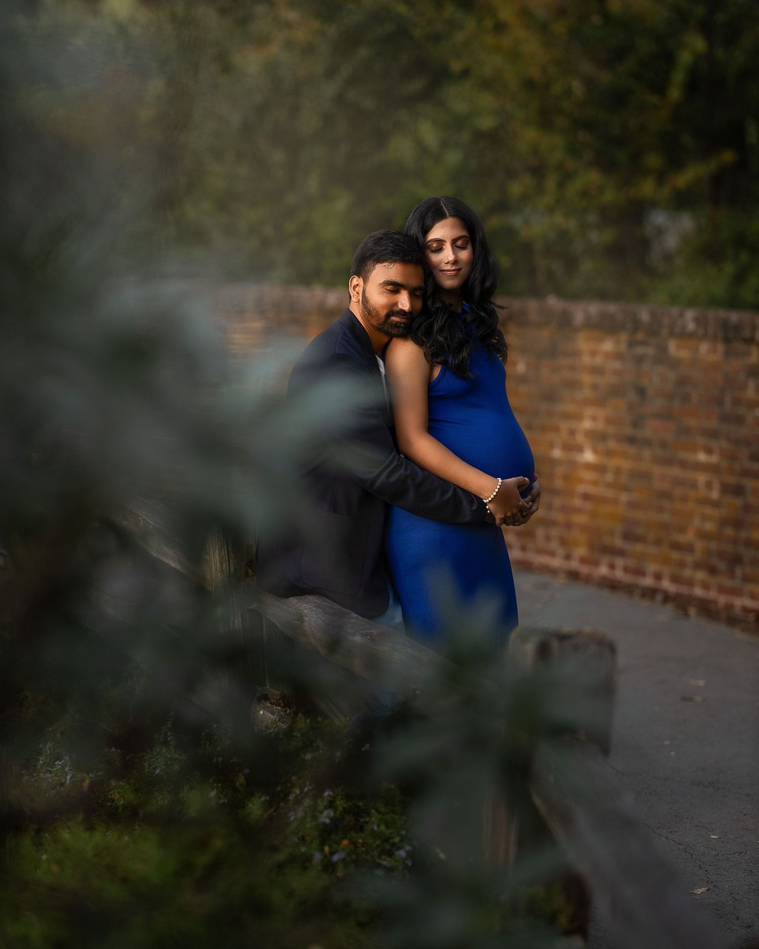 A couple embraces in front of a brick wall during a romantic outdoor photoshoot with moody lighting and foliage.