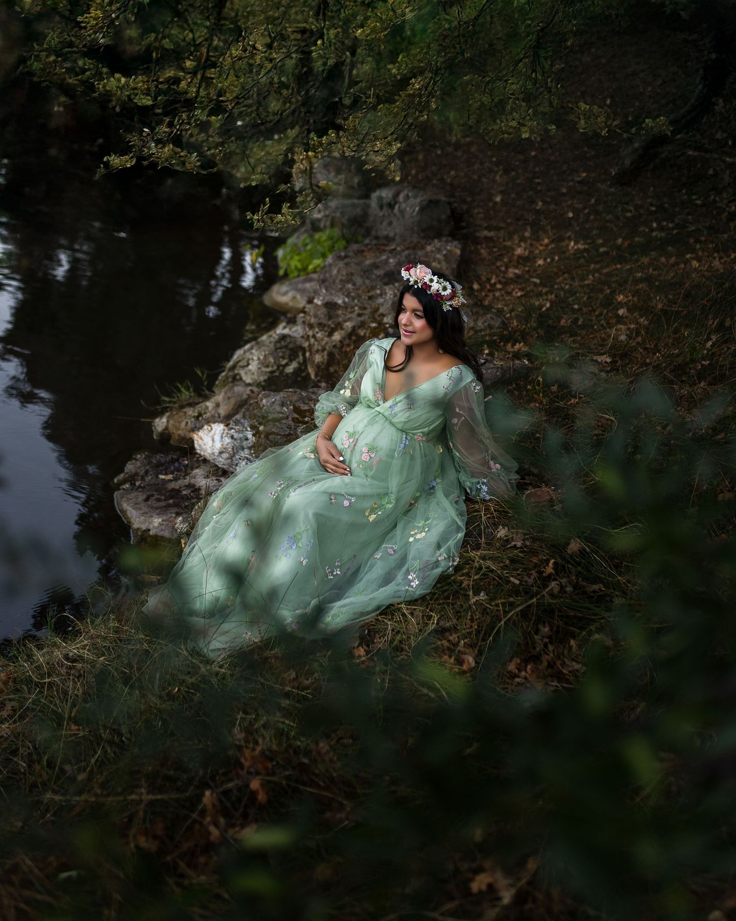 A person in a flowing mint green dress poses dramatically on rocks near dark water in a dreamy forest setting.
