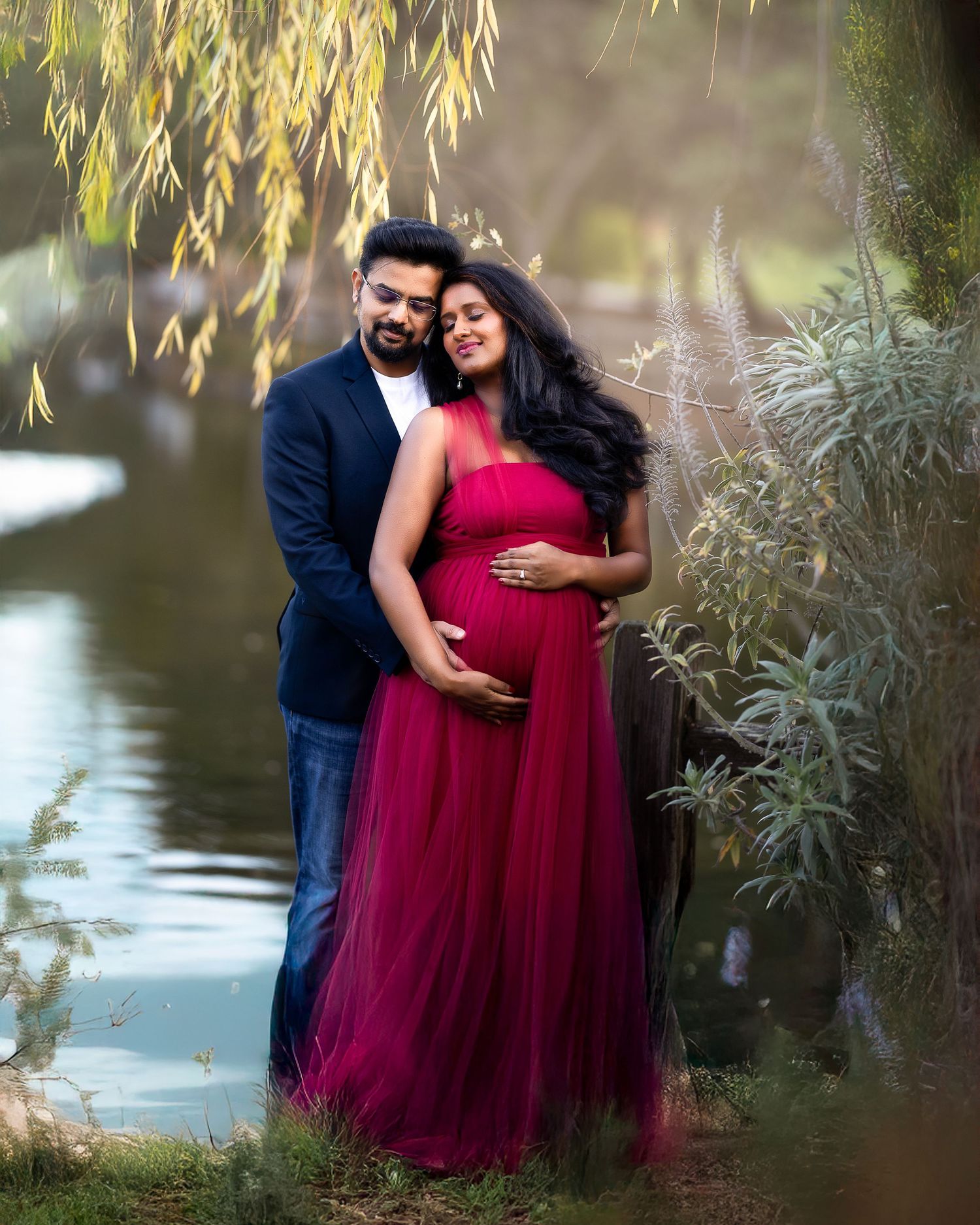 A couple in formal attire shares a romantic moment under willow trees by a tranquil pond.