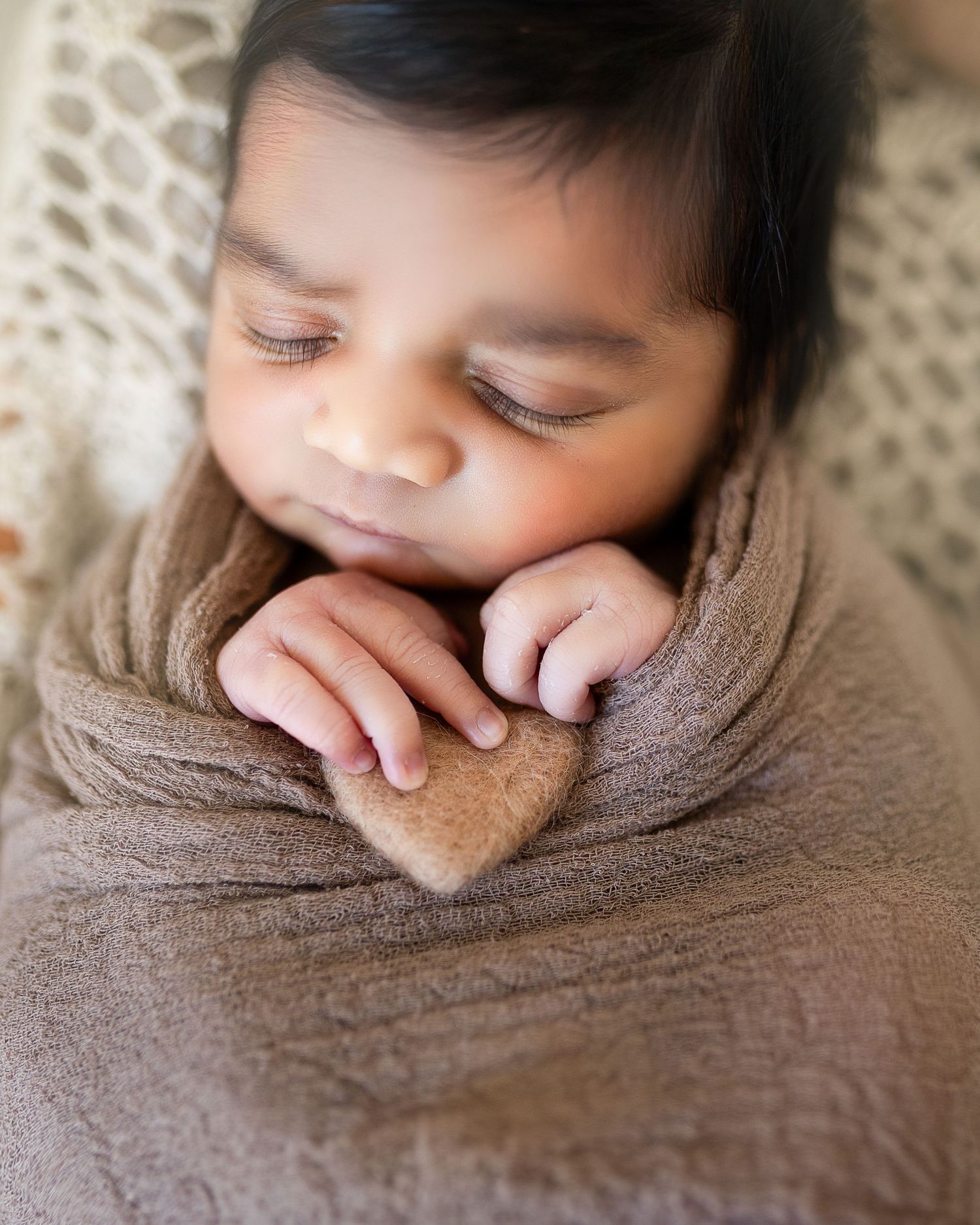 Close-up profile view of a sleeping newborn wrapped in brown fabric with tiny hands near face.