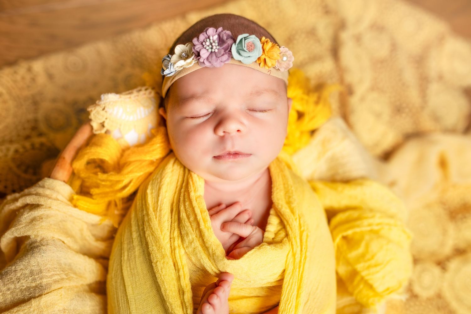 Newborn baby peacefully sleeping wrapped in soft yellow fabric with delicate floral headband.