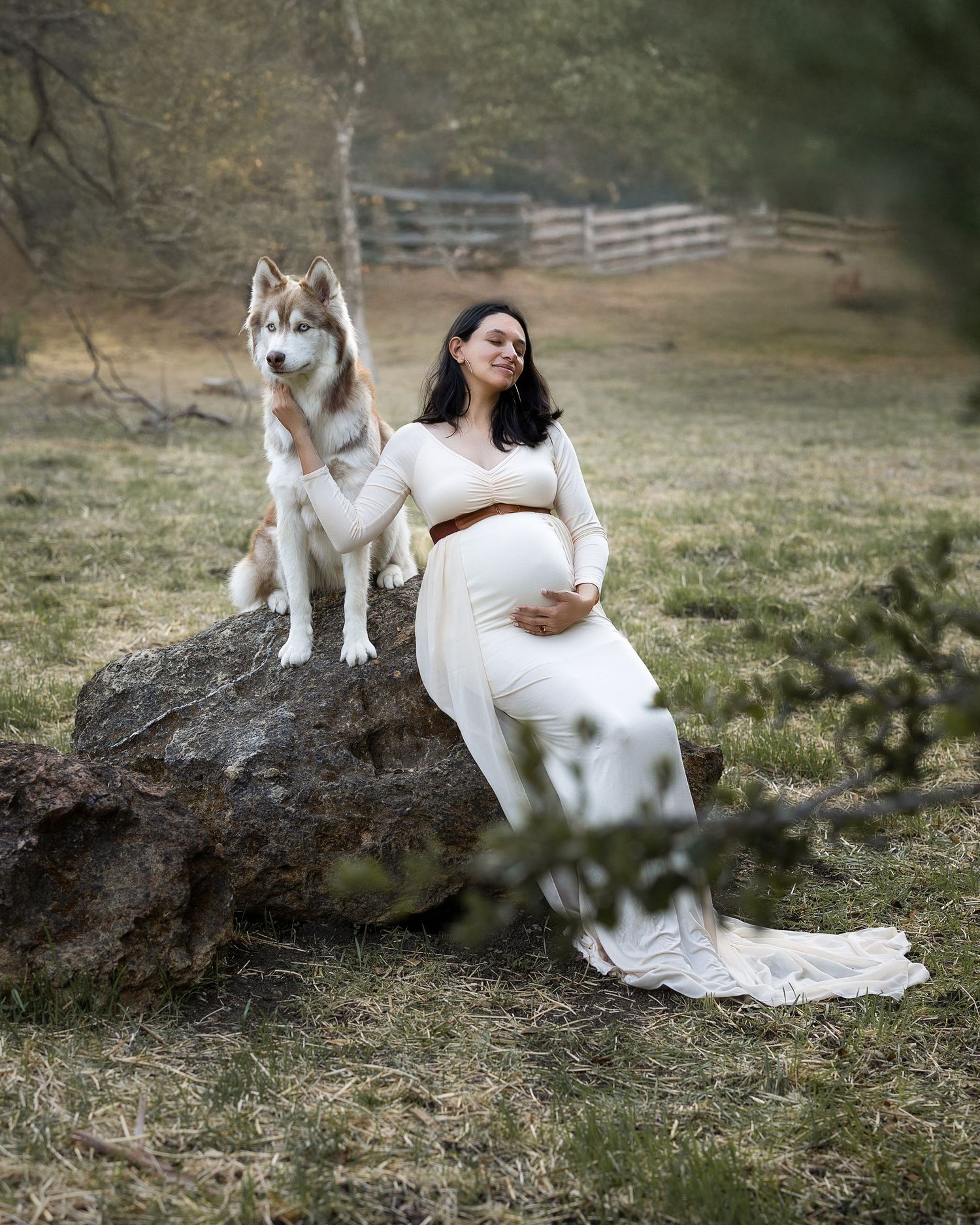 A person in a flowing white dress sits gracefully on a fallen tree trunk with a white cat in a natural outdoor setting.