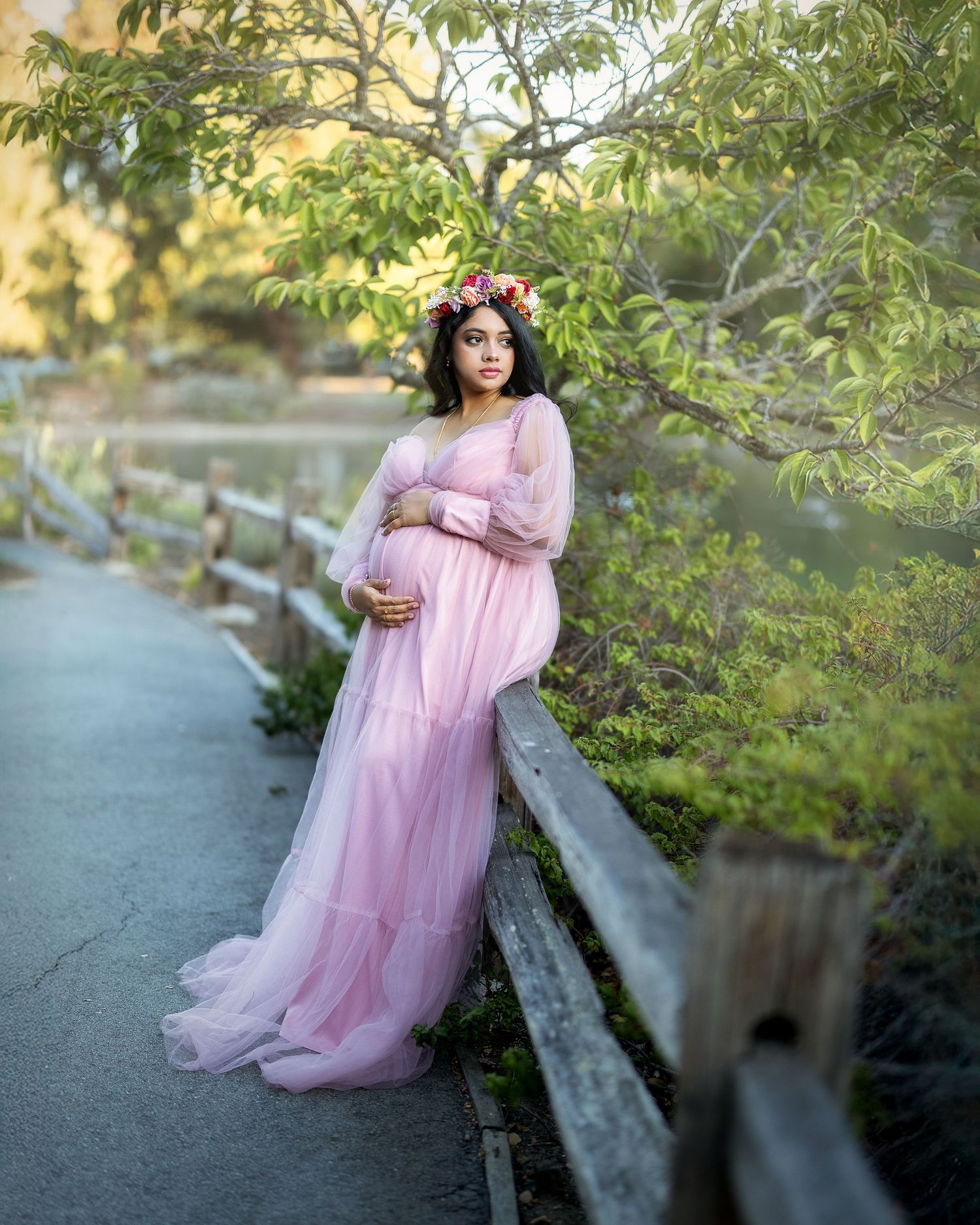 A pregnant woman in a flowing pink tulle gown poses gracefully in a natural outdoor setting during golden hour.