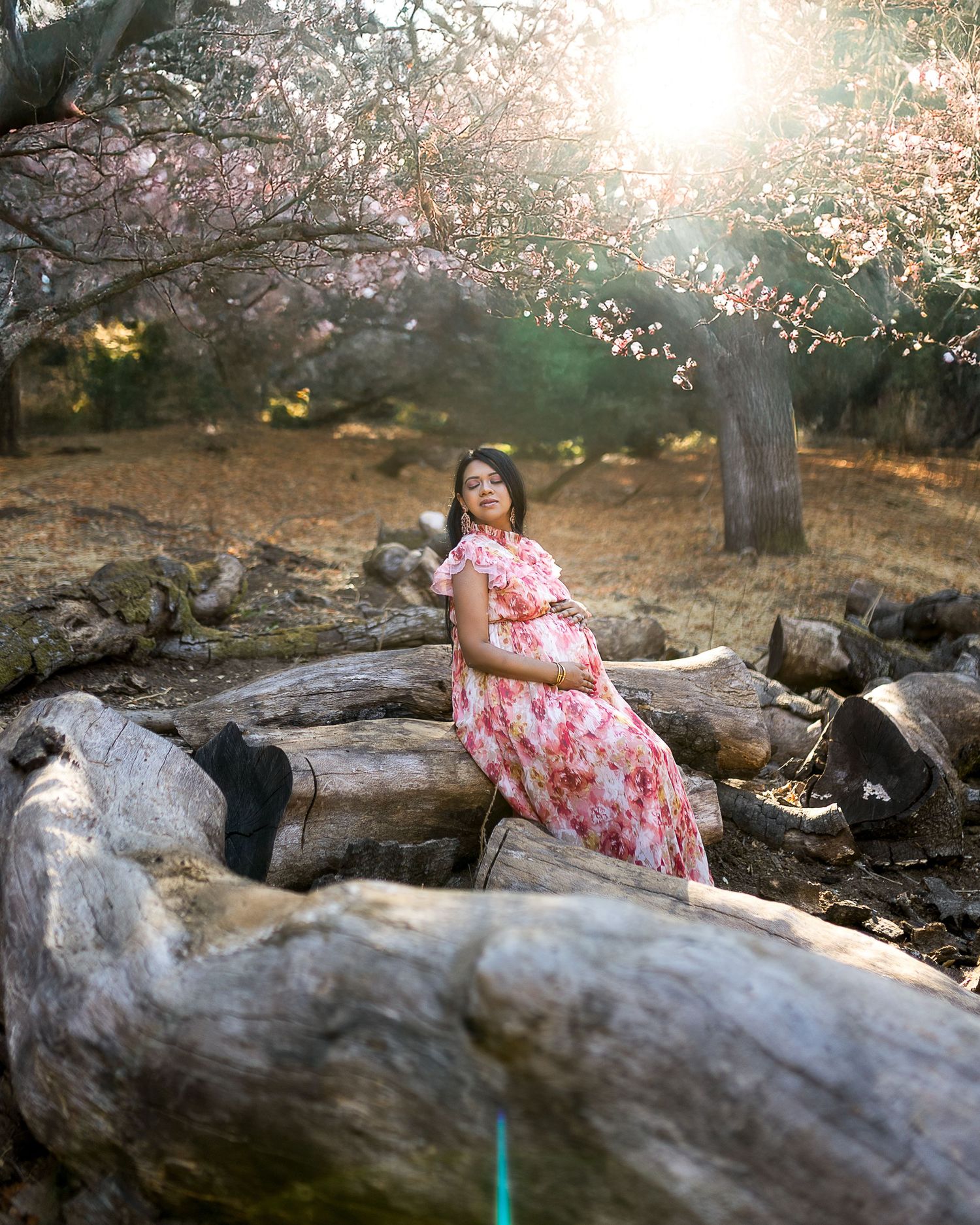 A person in a pink floral dress poses on a fallen log in a dreamy outdoor setting with sunlit cherry blossoms.