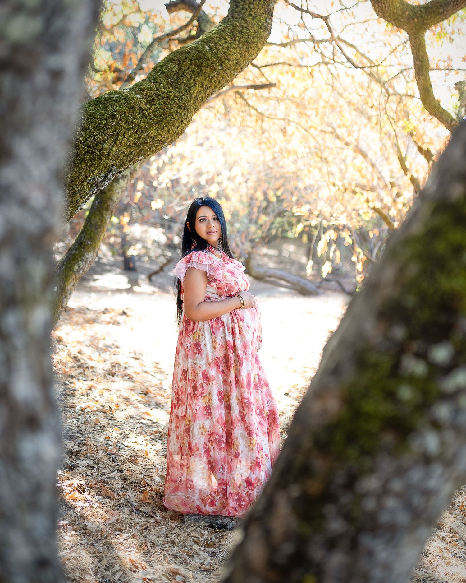 A figure in a pink floral dress stands beneath blooming cherry blossom trees on a sunny spring day.