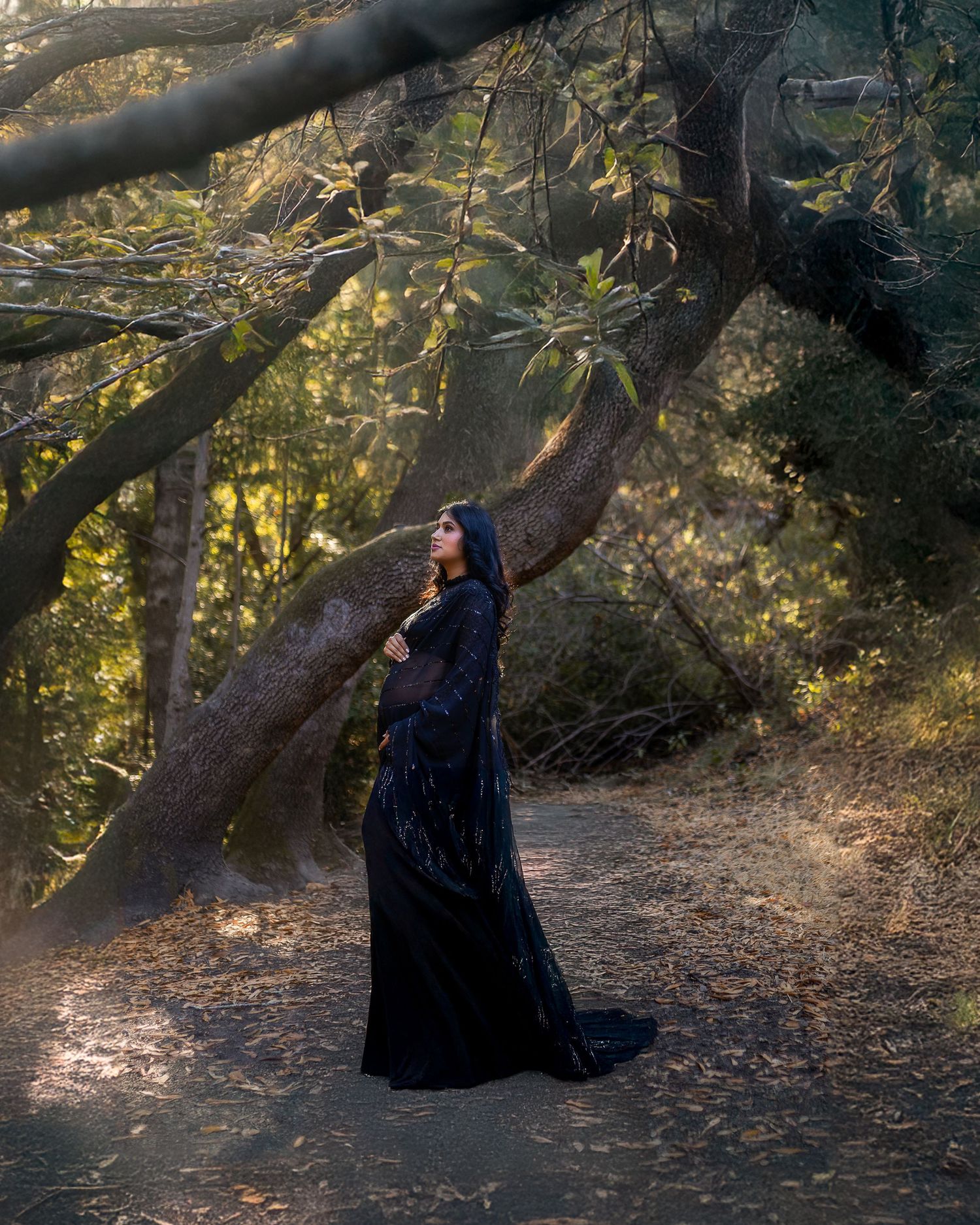 Mysterious figure in flowing black dress poses dramatically under sunlit tree branches in a forest setting.