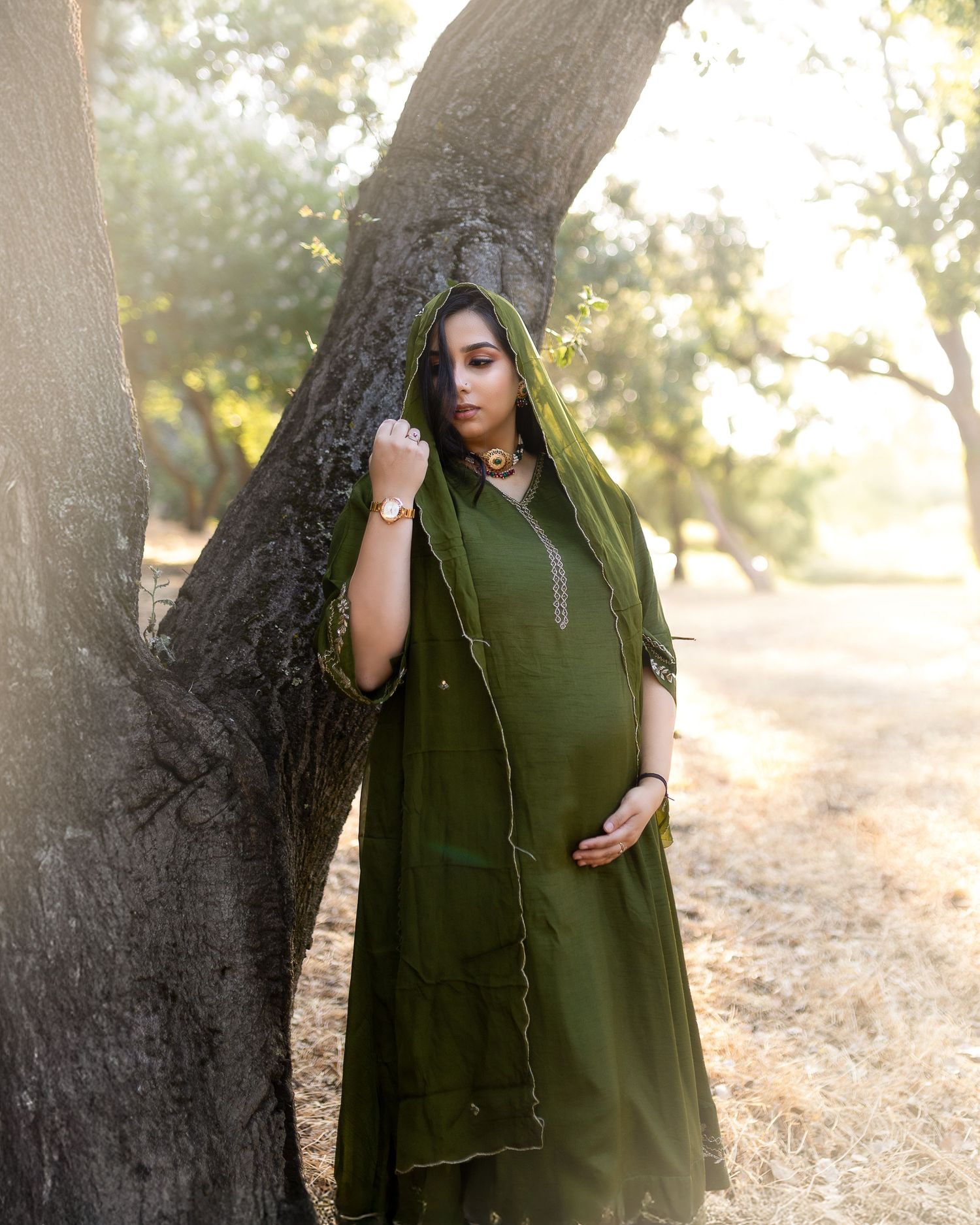 A figure in a dark olive green cloak stands mysteriously among trees in soft natural lighting.
