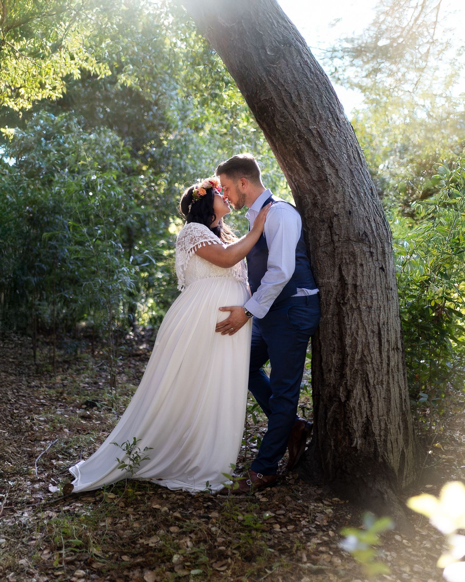 A romantic wedding couple shares intimate moments under a large tree in a sunlit outdoor setting.