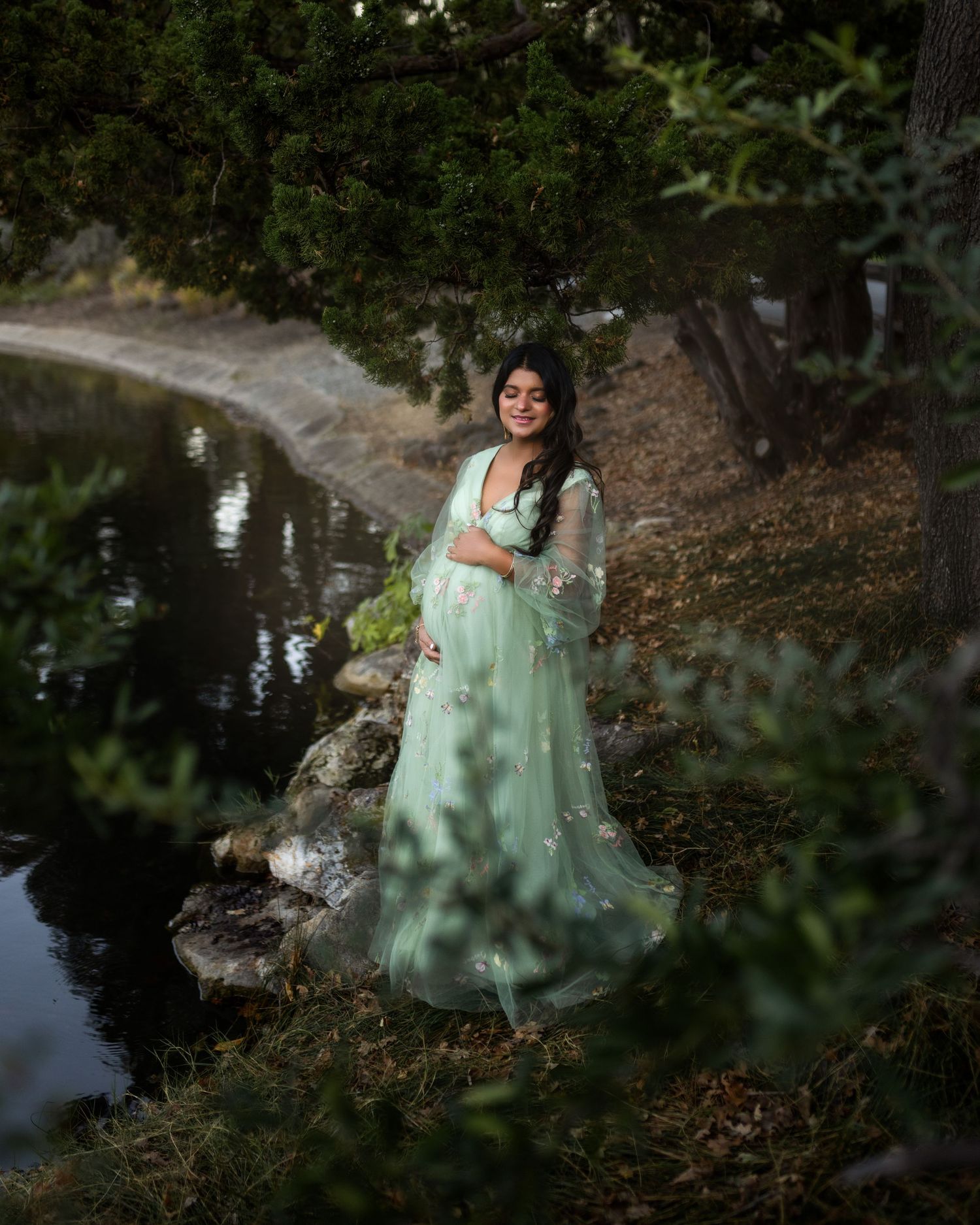 A person in a mint green flowing dress poses gracefully by a reflective pond in a wooded garden setting.