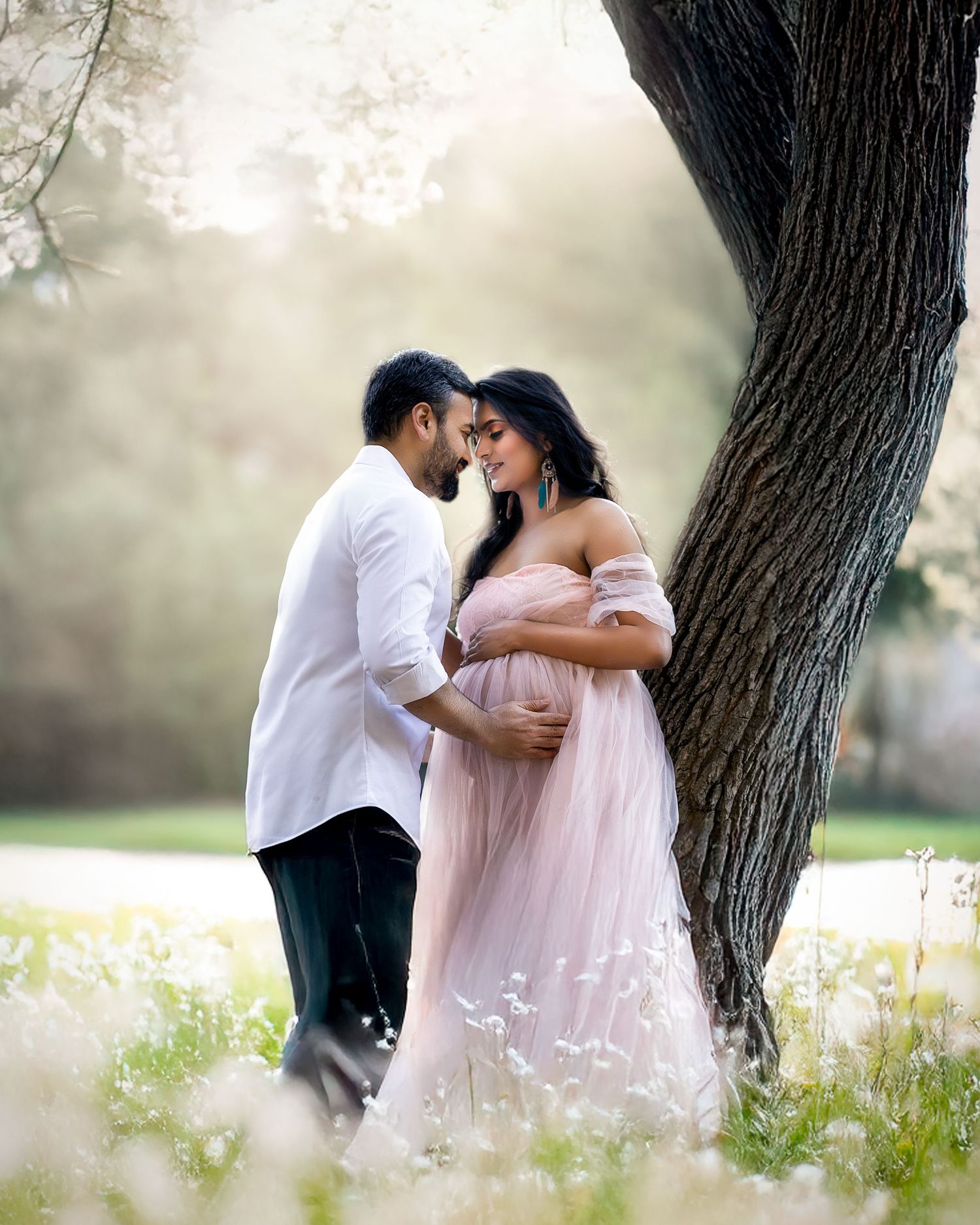 A couple in white and pink outfits embrace under a large tree in a dreamy outdoor setting with wildflowers.