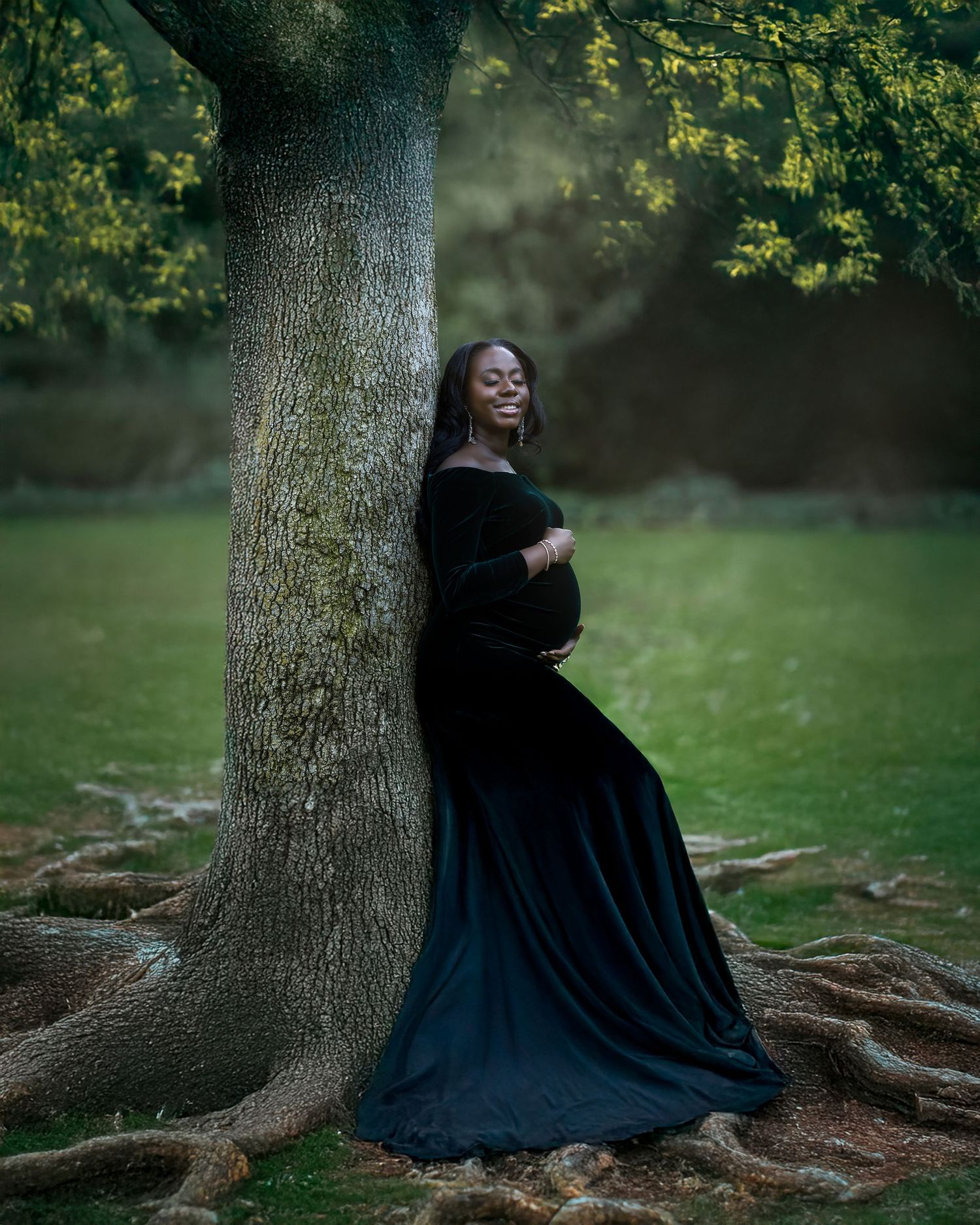 Person in flowing black dress sits gracefully against large tree trunk in moody outdoor setting.