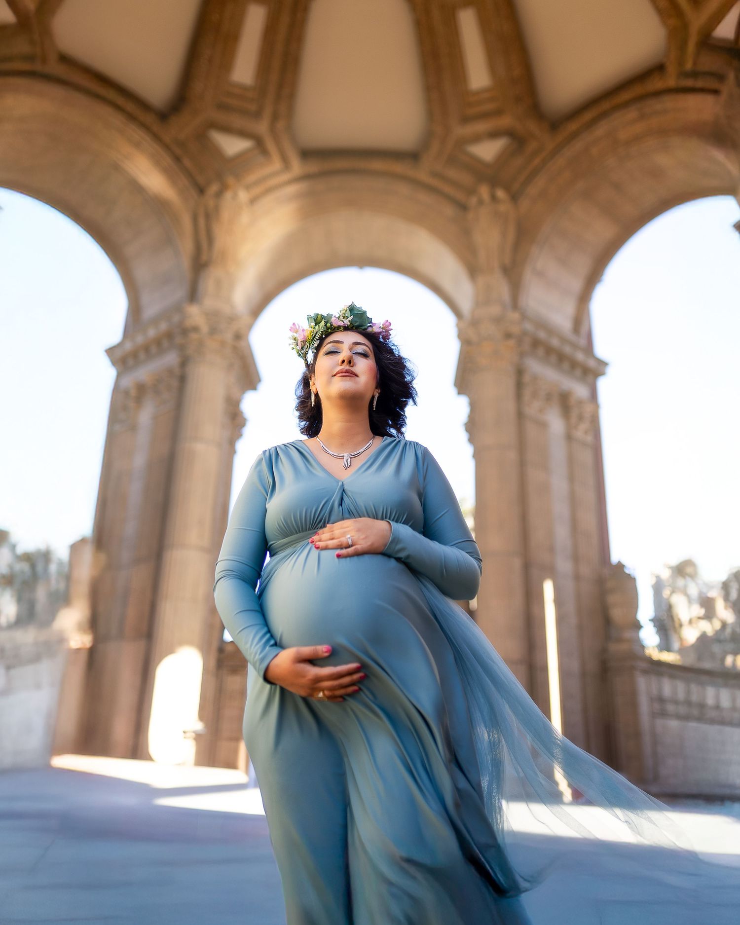 Maternity photoshoot in a light-filled stone archway corridor featuring a woman in a flowing blue dress.