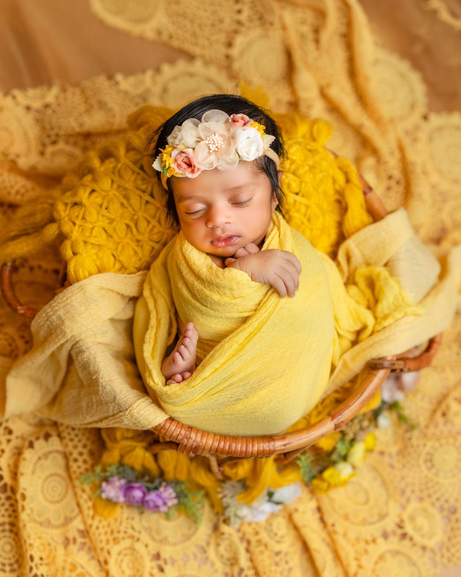Newborn baby wrapped in yellow swaddle blanket sleeping peacefully on textured cream backdrop with floral accents.