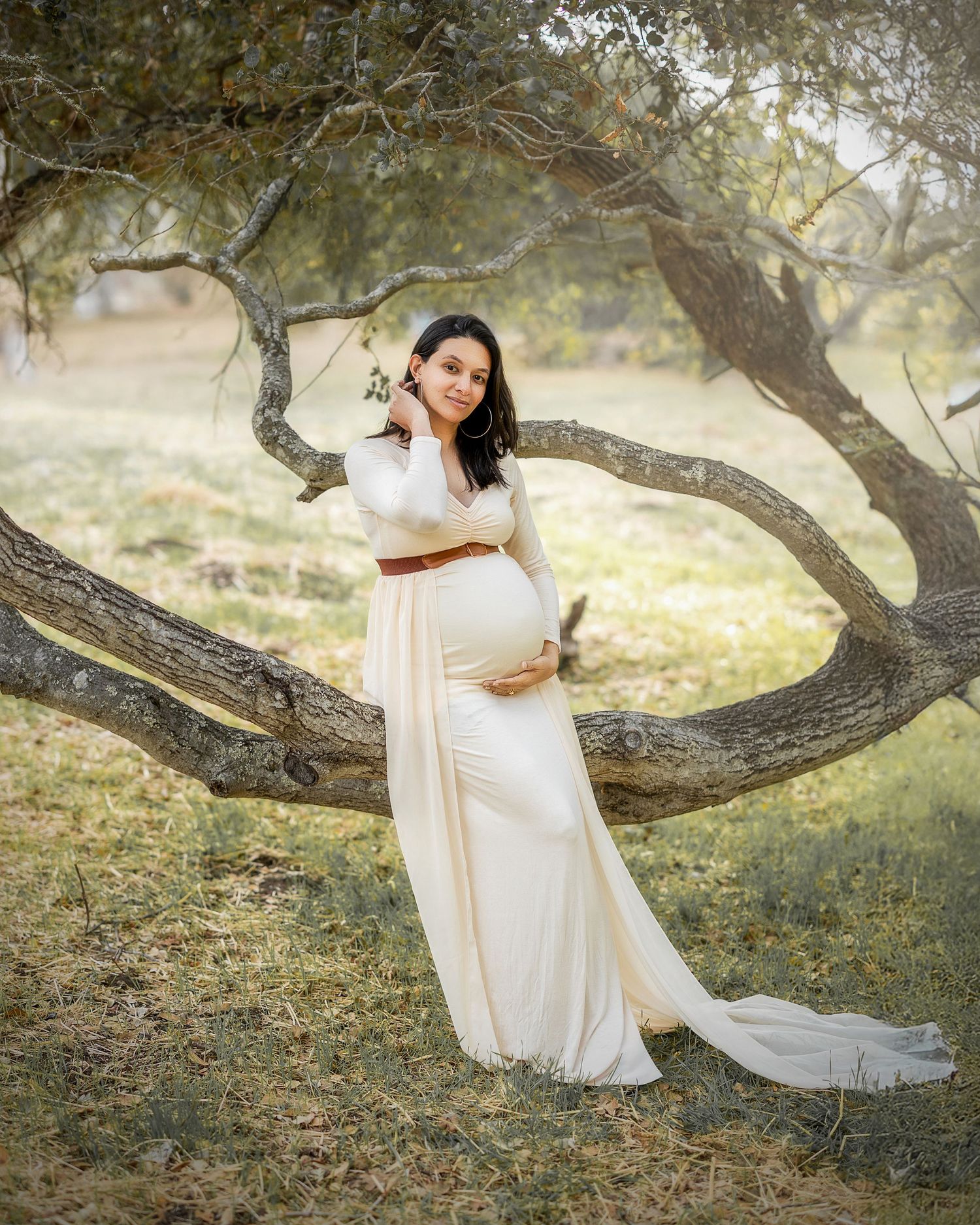 Expectant mother in flowing white dress poses gracefully against twisted tree branches in a dreamy outdoor setting.