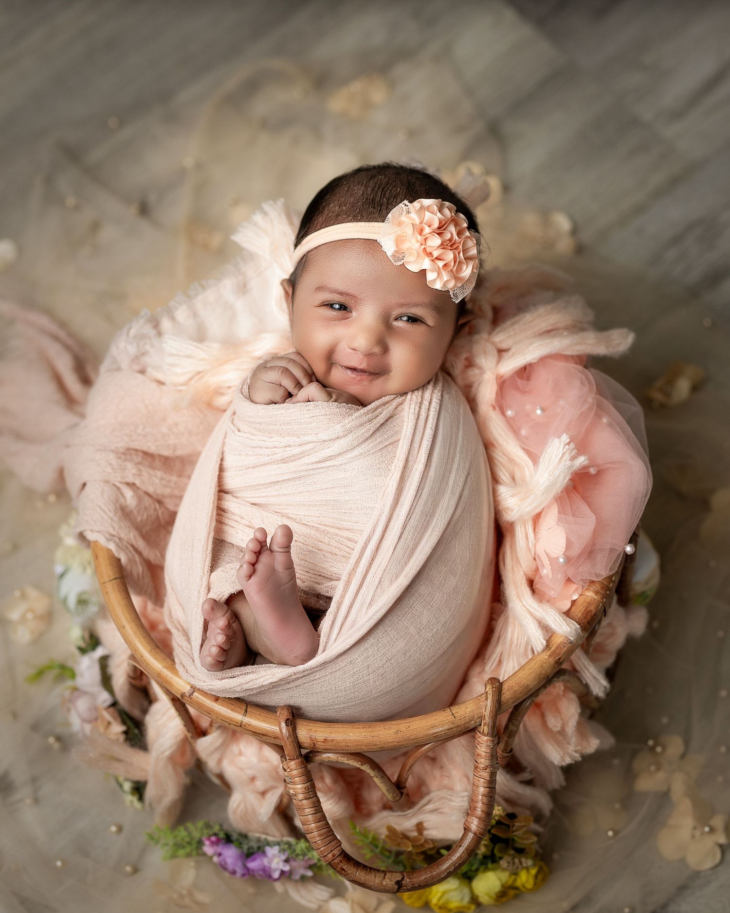 A baby wrapped in soft beige fabric lies in a rustic basket surrounded by peach-colored floral decorations.