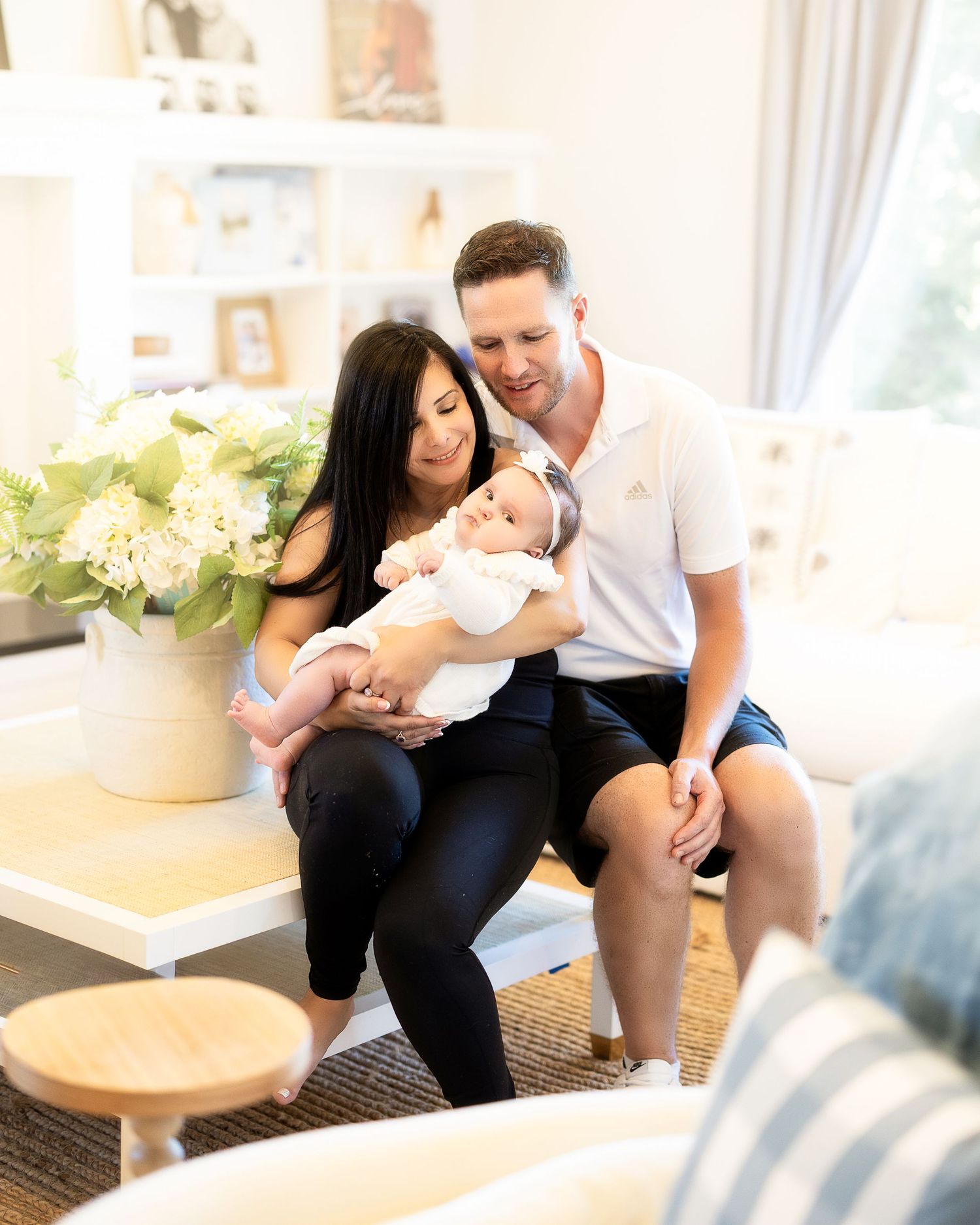 A couple sits together on a couch with a baby while a vase of white hydrangeas decorates the bright interior space.
