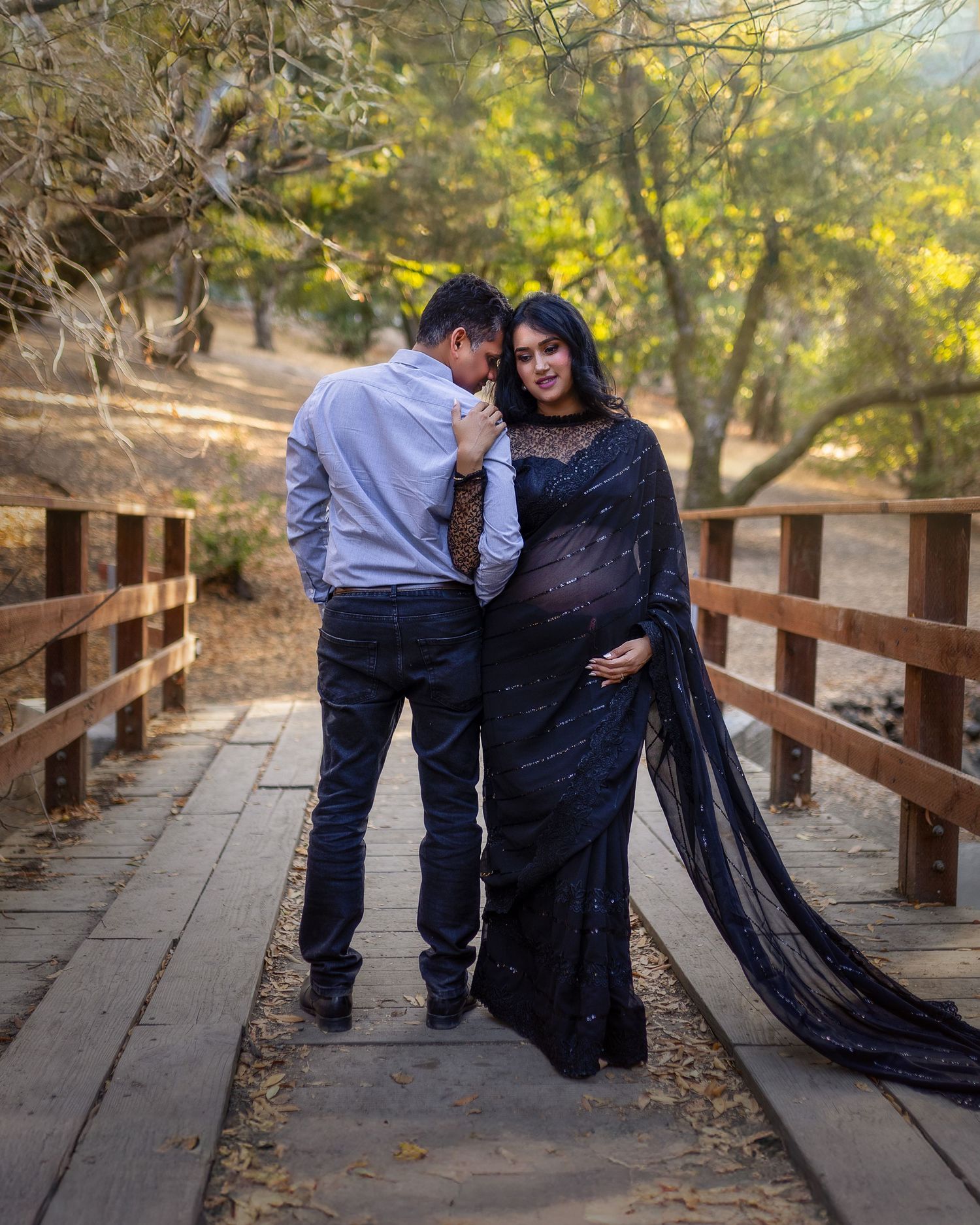 A couple embraces on a wooden bridge pathway surrounded by autumn trees with golden sunlight filtering through.