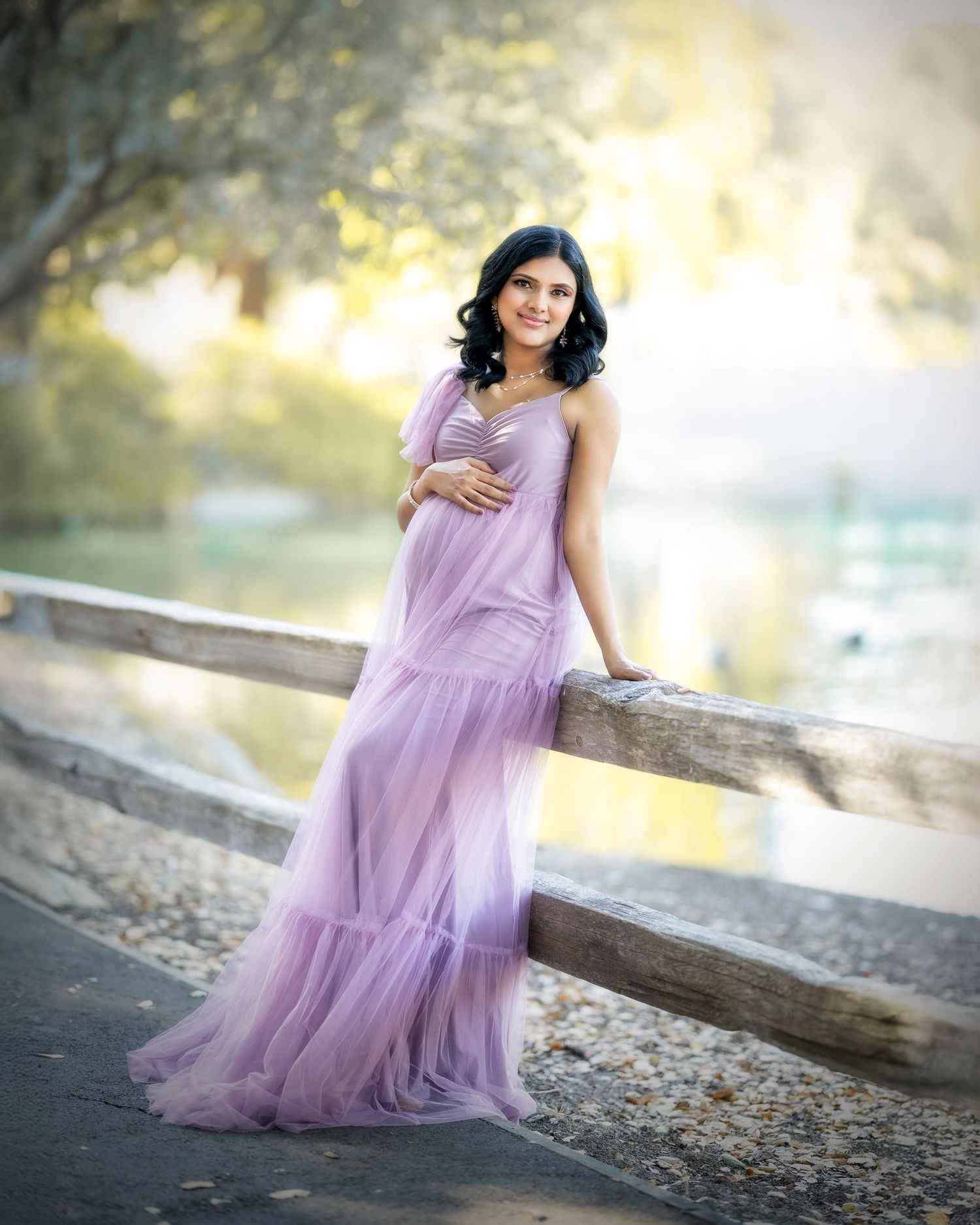 Dreamy outdoor maternity photos featuring a flowing purple tulle gown by a waterfront with golden sunlight.