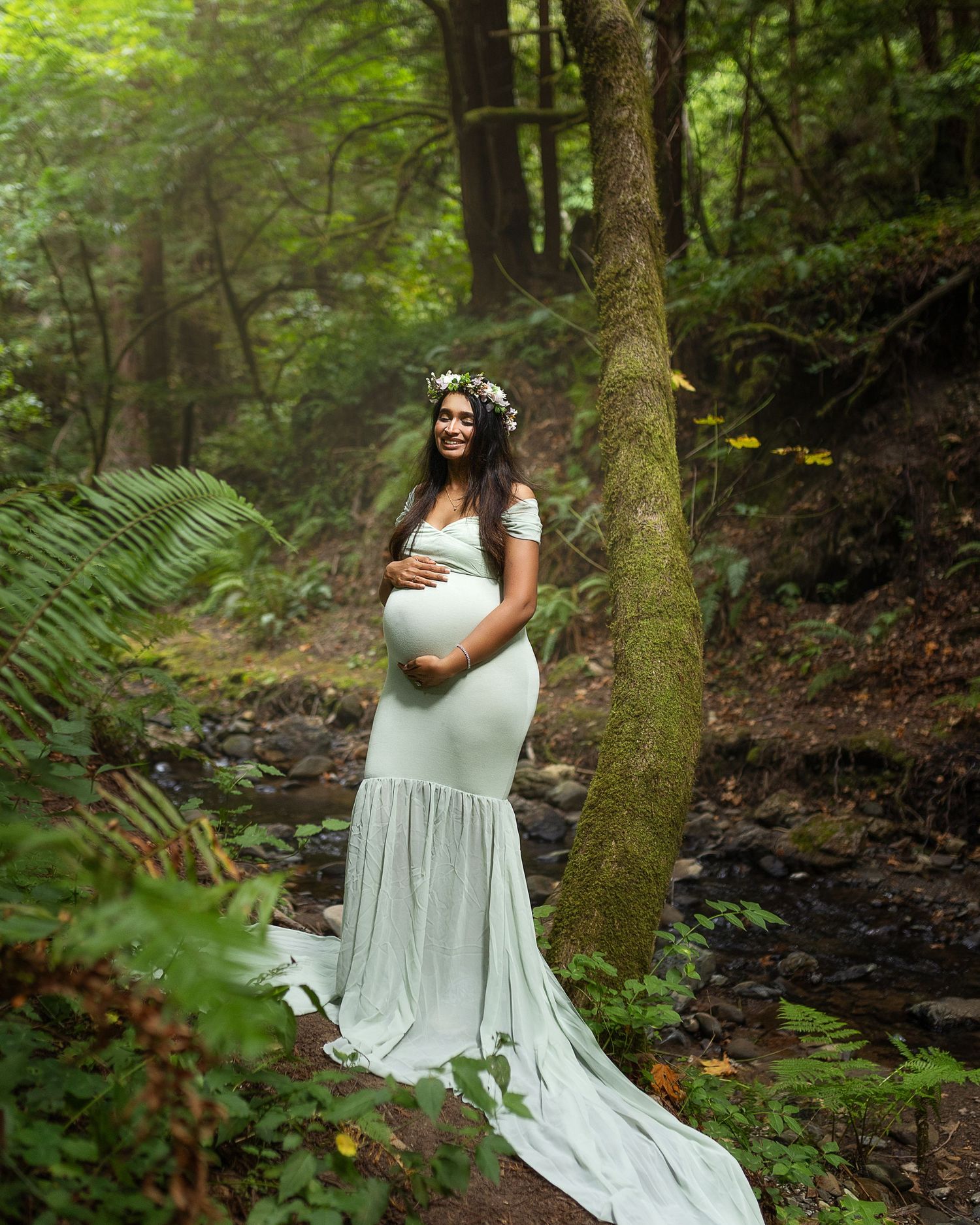 A pregnant woman in a flowing white dress poses gracefully in a lush green forest setting near a stream.