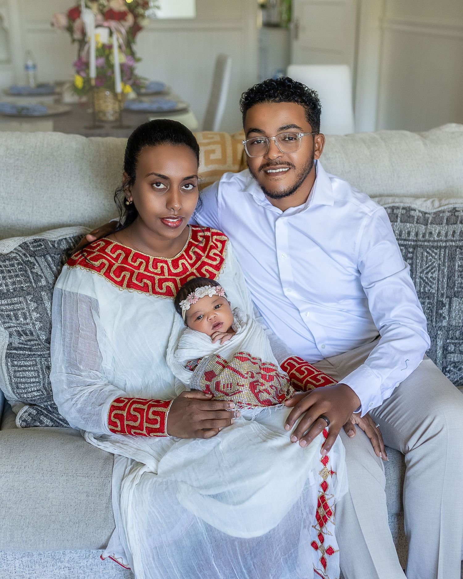 A family relaxes together on a gray couch wearing coordinated white and red traditional Ethiopian outfits.