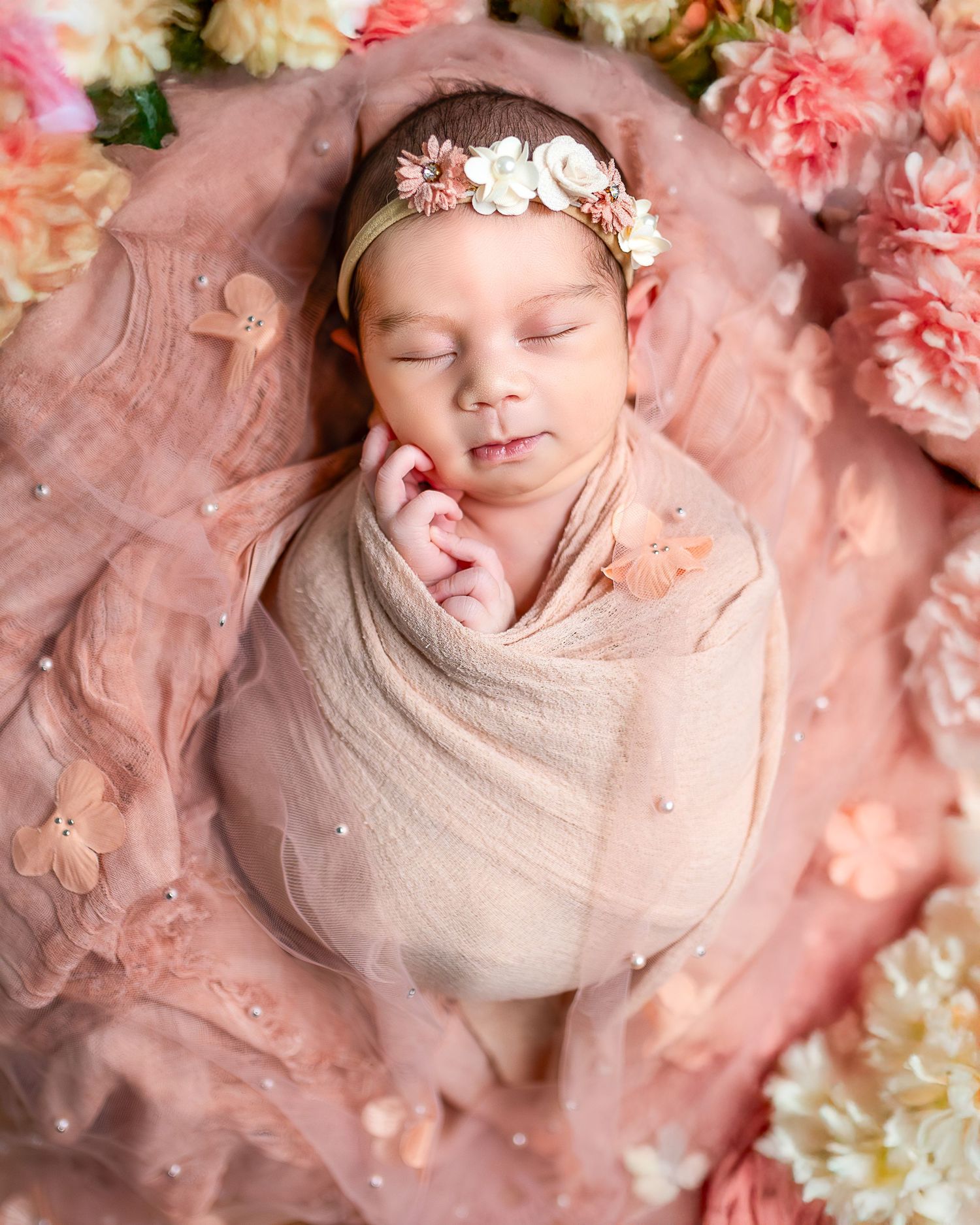 Sleeping newborn wrapped in beige swaddle on pink fabric surrounded by pink and cream flowers.