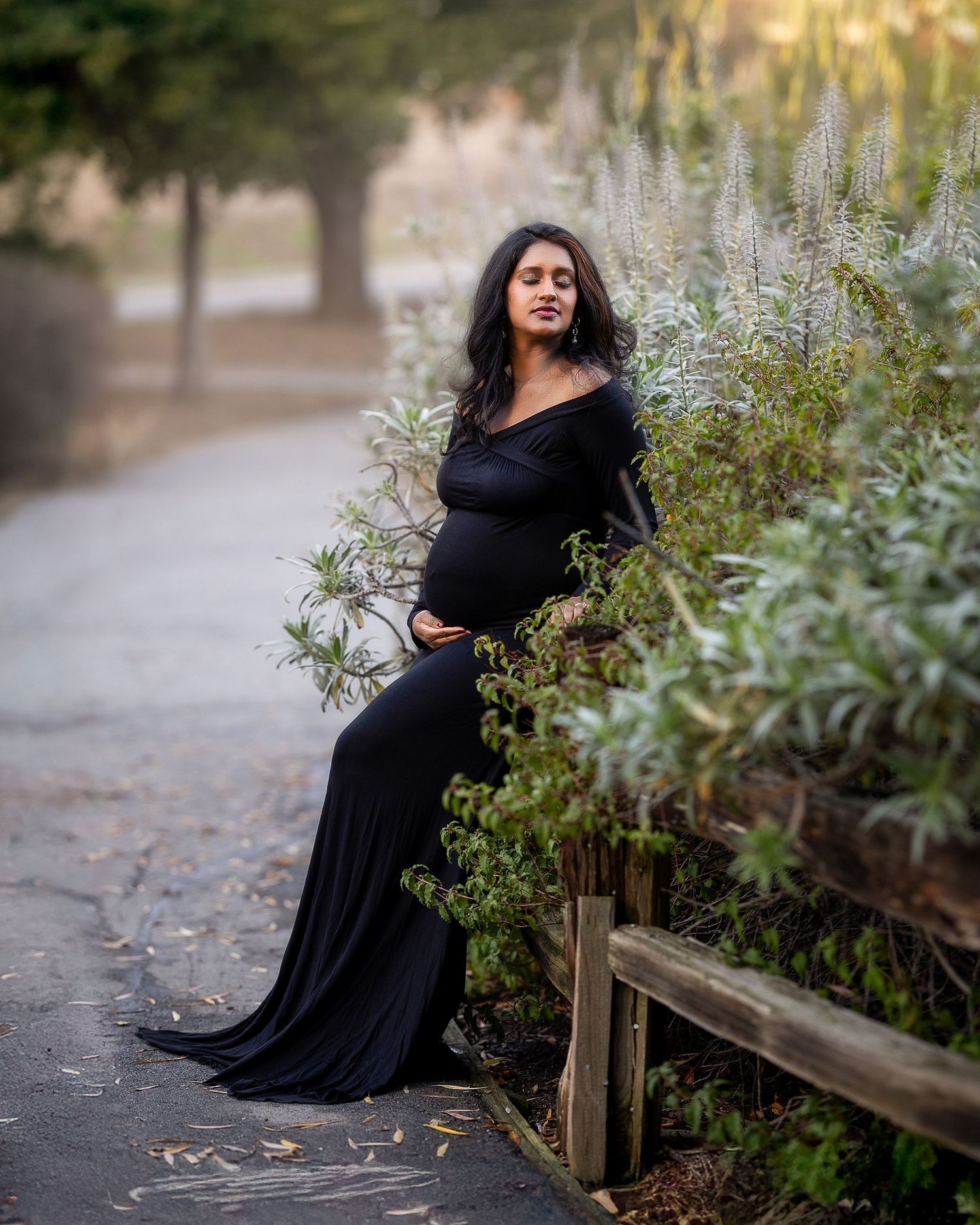 A figure in a flowing black dress poses gracefully in a garden pathway during golden hour lighting.