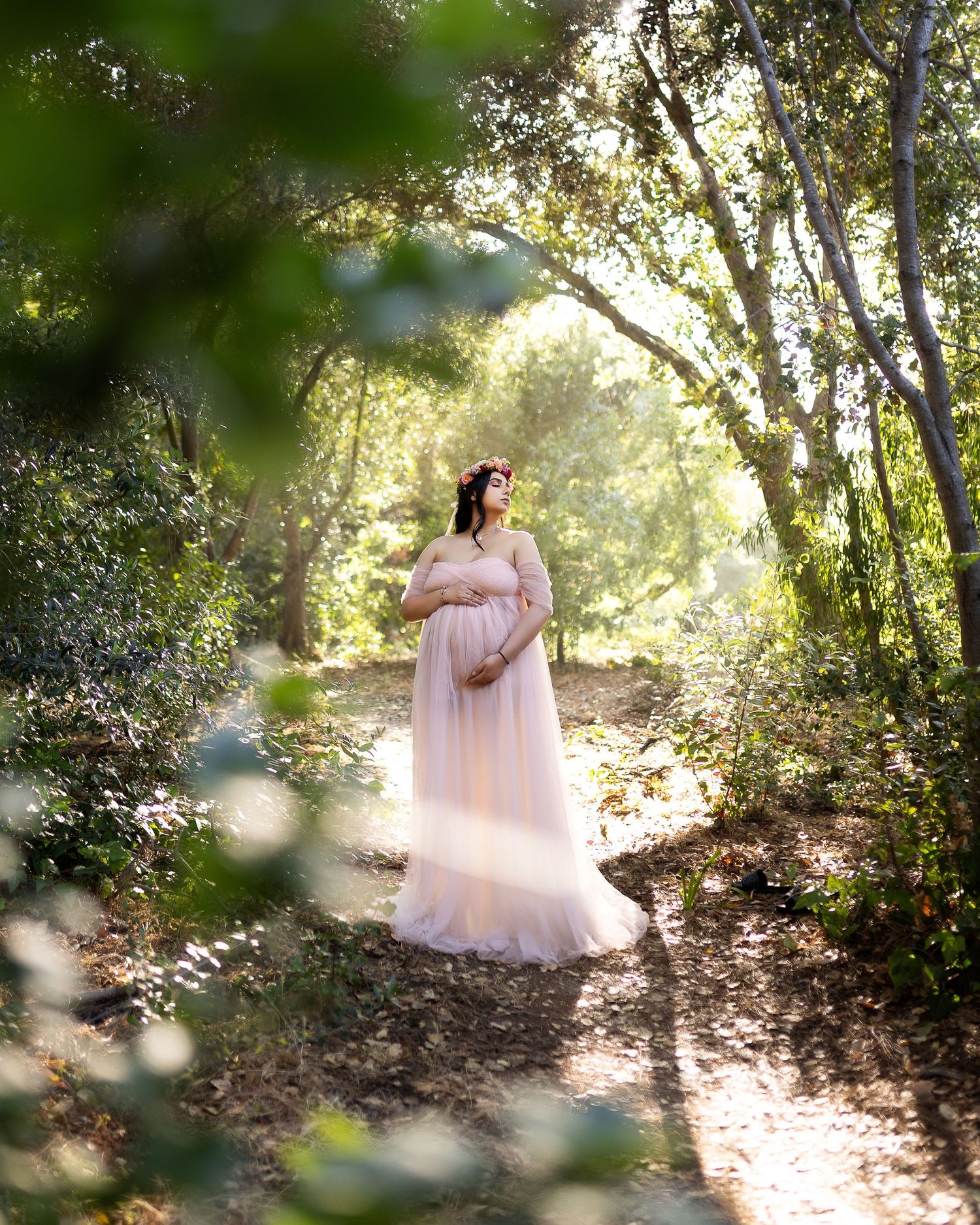 A dreamy maternity photo series shows a woman in a flowing pink gown walking through a sunlit wooded forest path.