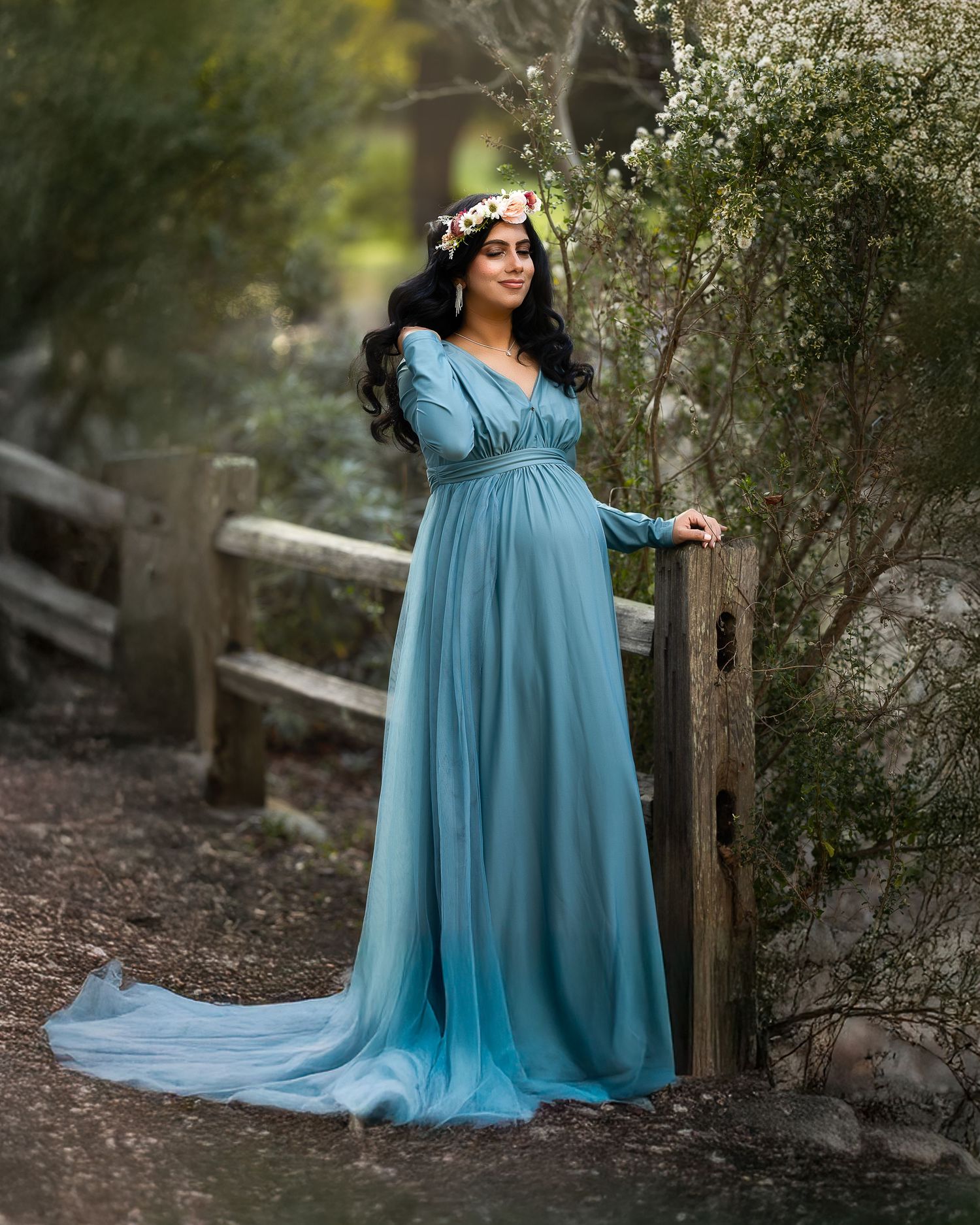 A person in a flowing light blue maternity gown poses gracefully in a wooded outdoor setting during a photo session.