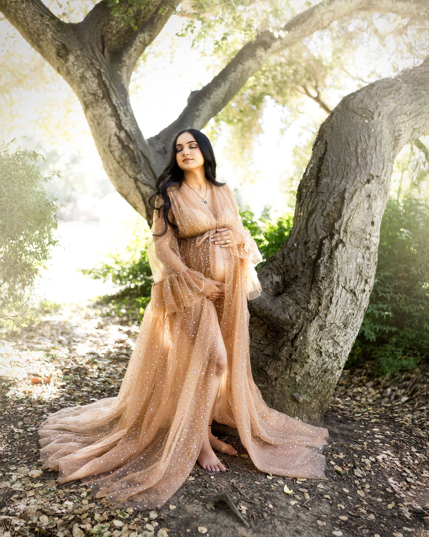 Ethereal maternity photo featuring a flowing champagne-colored gown with glitter details against large tree trunks in soft light.