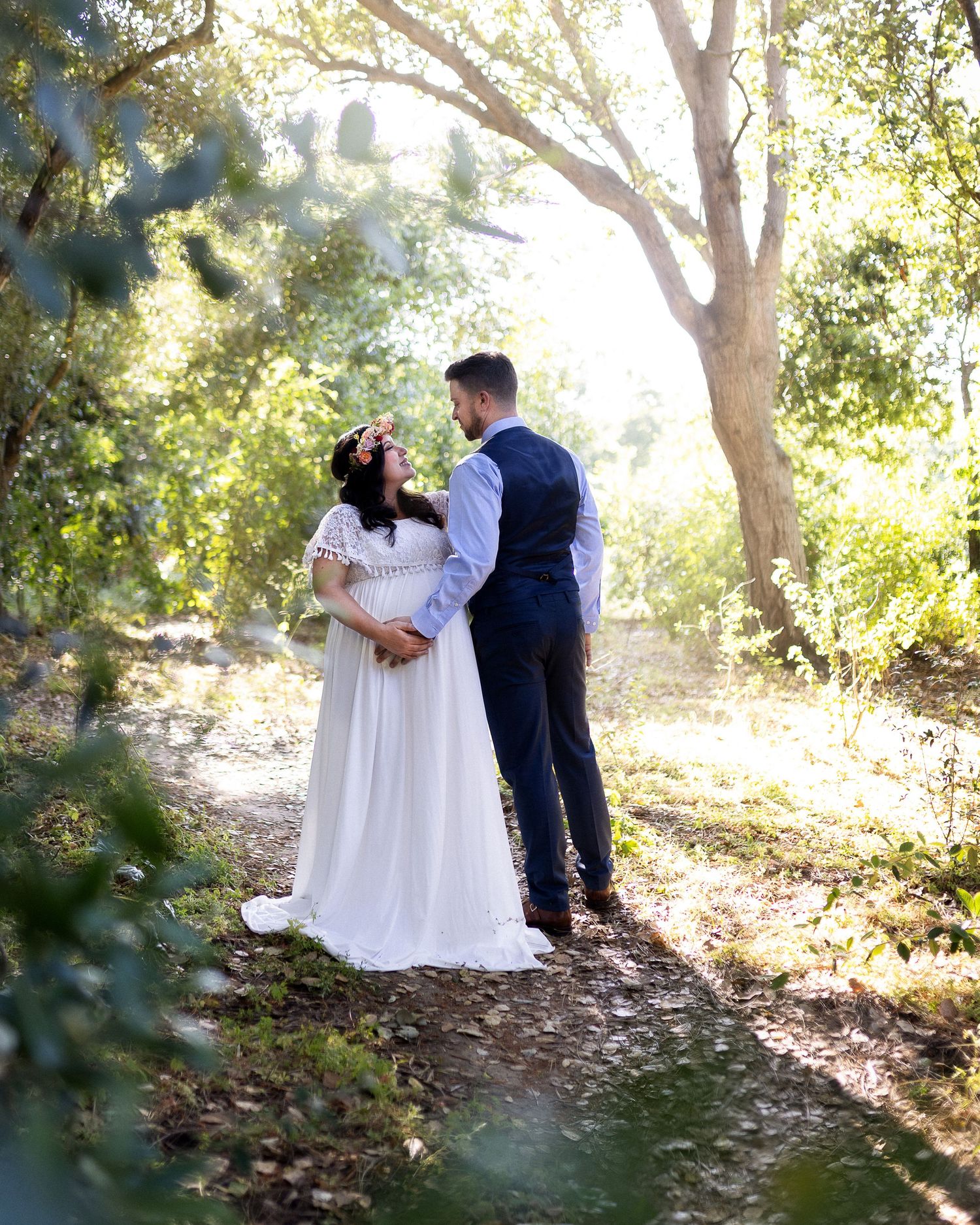 A romantic outdoor wedding photoshoot with a couple in white dress and navy suit dancing among sunlit trees.
